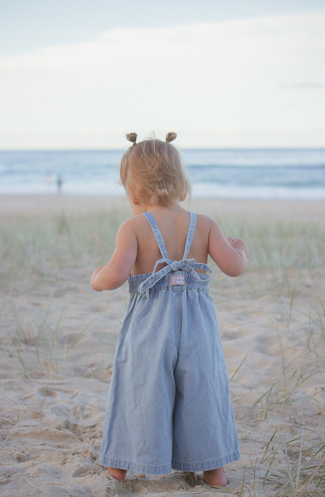 A toddler with two small pigtails stands facing the ocean on a sandy beach, wearing the Jane Jumpsuit by TWIN COLLECTIVE. The child is surrounded by patches of grass and appears to be looking at the calm water under a lightly clouded sky.