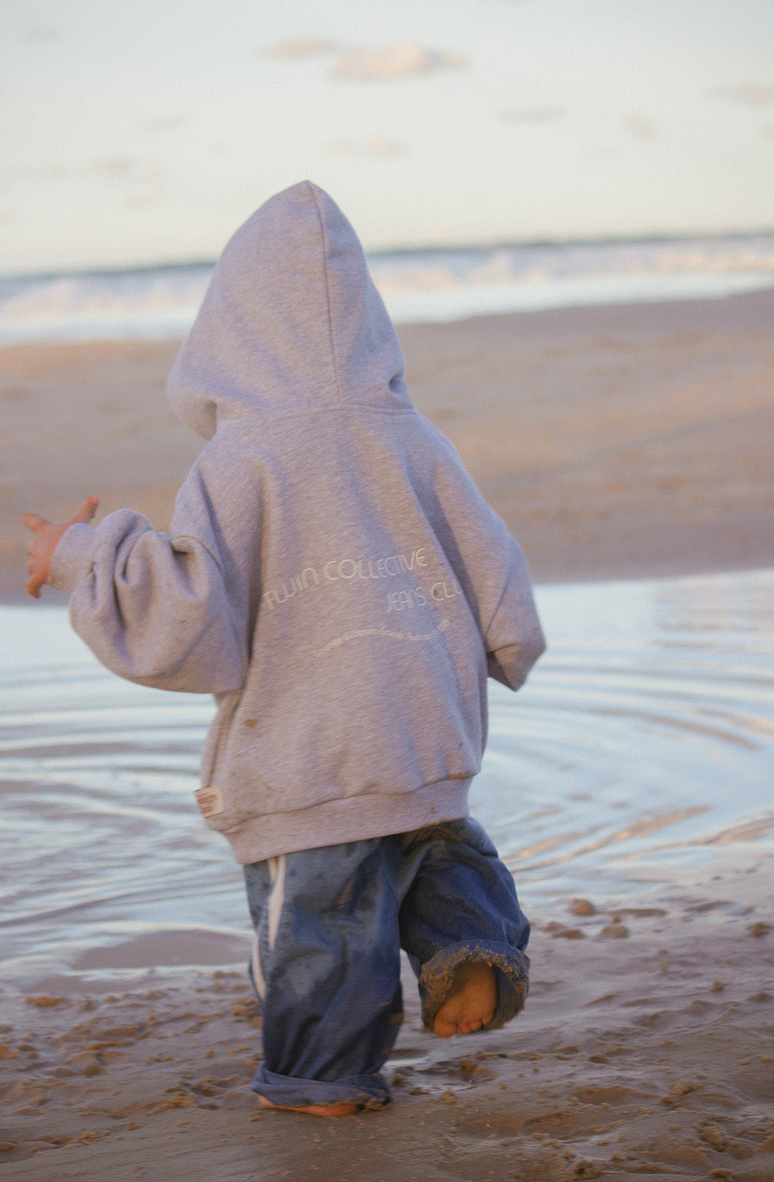 A young child wearing an oversized TWIN COLLECTIVE Logo Hoodie Set Grey Marle and baggy jeans walks barefoot along a sandy beach. The child is facing away, with the large hood covering their head. They are near a shallow pool of water, with the ocean and sky visible in the background.