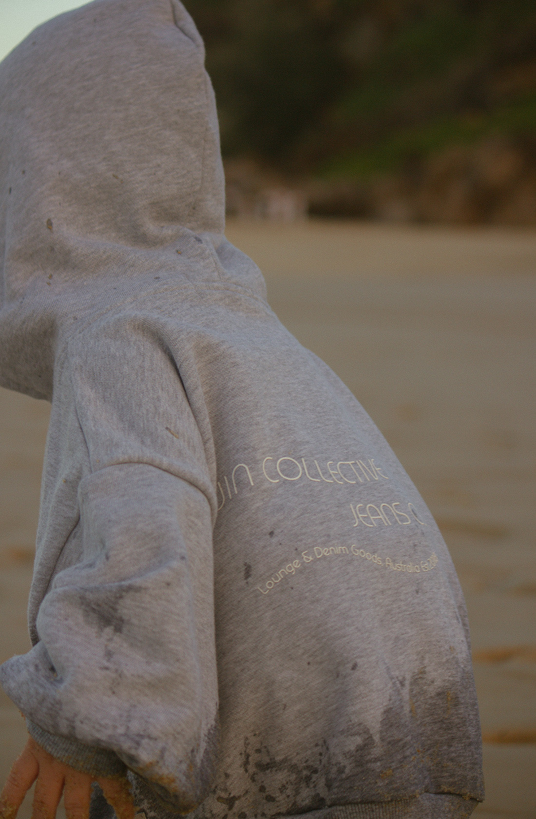 A child wearing the Logo Hoodie Set Grey Marle from TWIN COLLECTIVE, with partially obscured text on the back, plays in a sandy area that seems like a beach. The hoodie, crafted from cozy Loopback Terry fleece, has some dirt or sand marks. Paired with oversized track pants, the background reveals blurred greenery and possibly a rock or hillside.