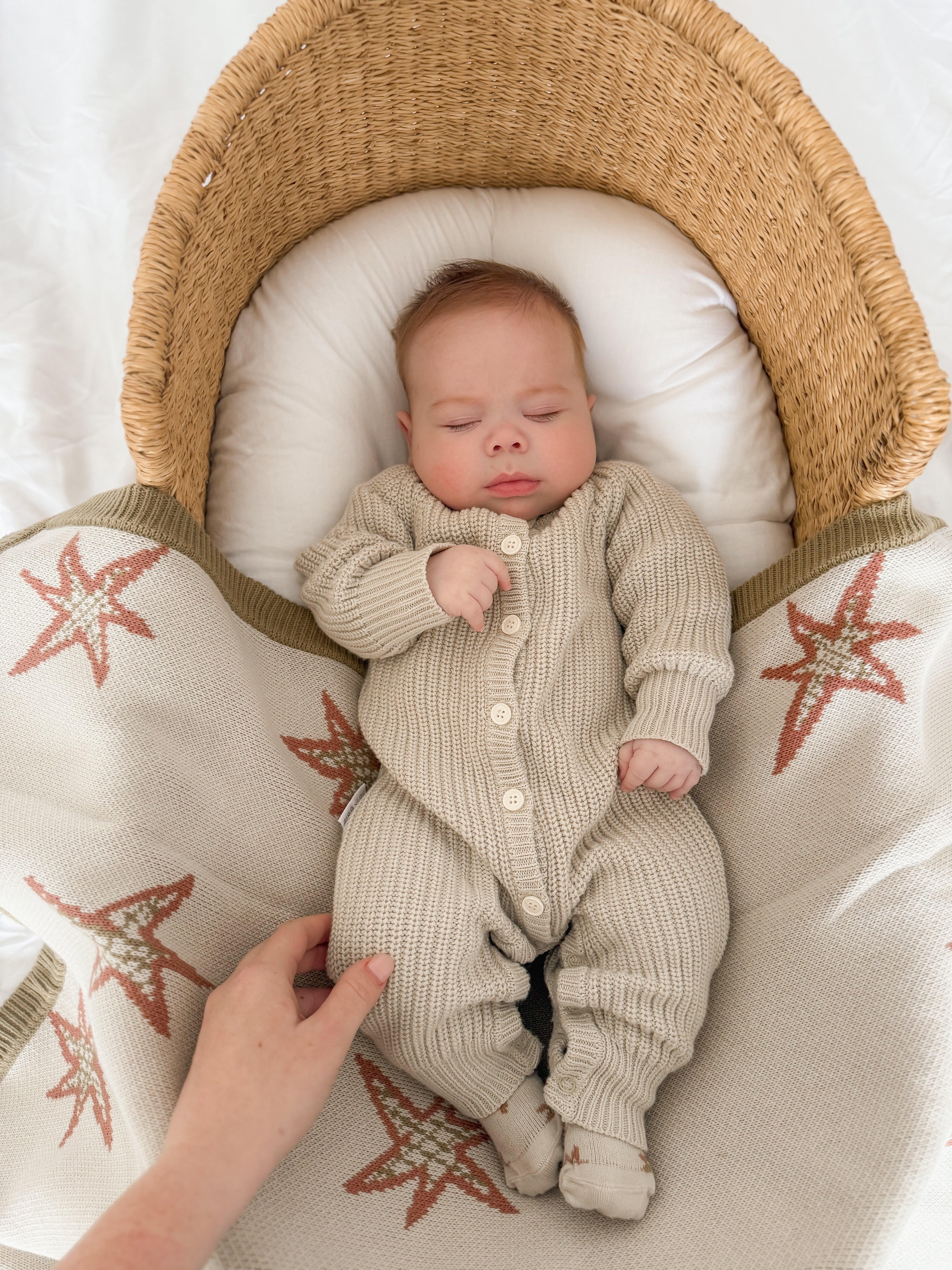 A baby wearing the ZIGGY LOU Ziggy Lou - Classic Knit Romper Moss sleeps peacefully in a wicker bassinet under a cream blanket with red and green star patterns, as an adult's hand gently rests on the blanket beside them.
