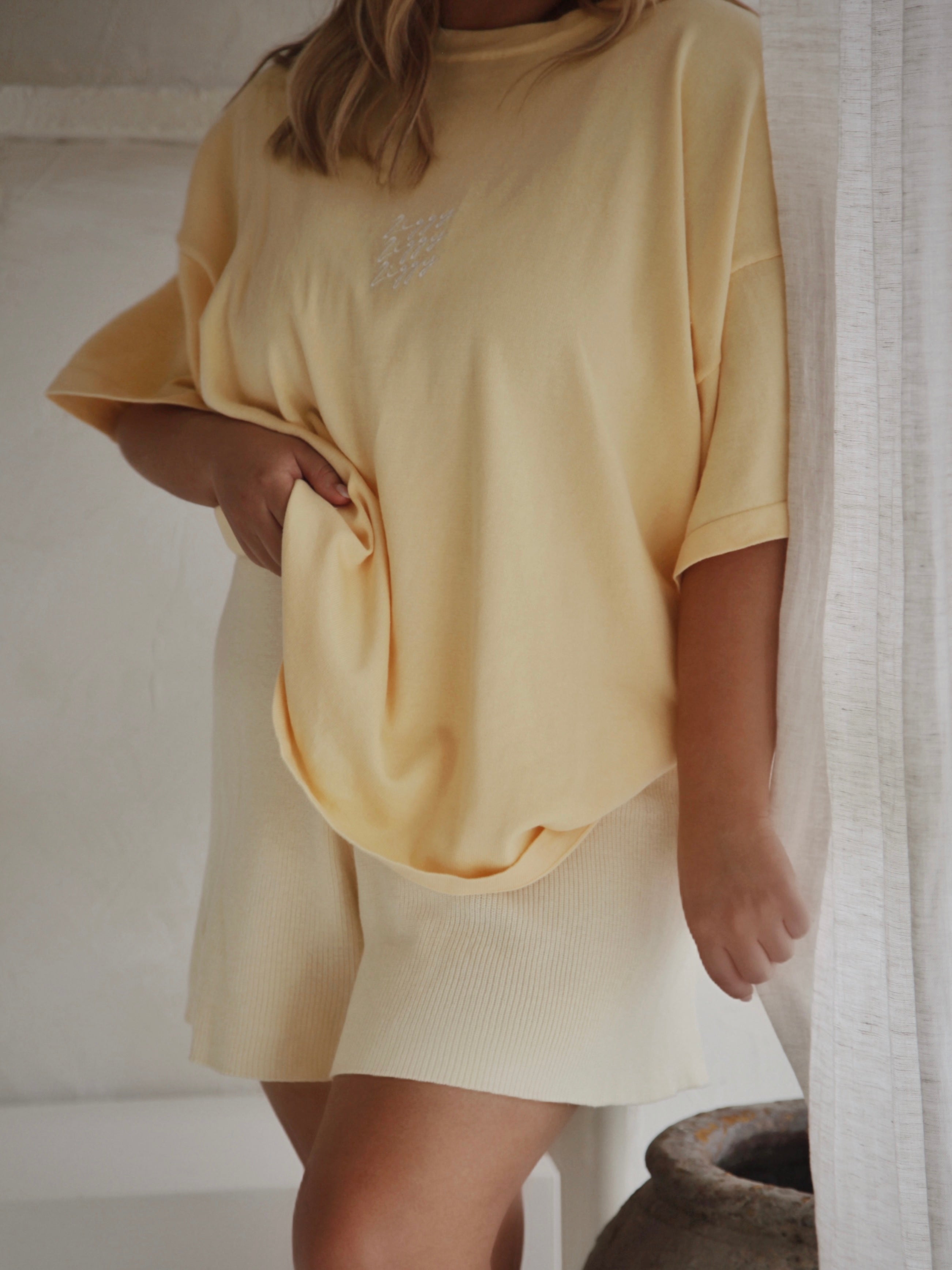 A person wearing the Women's Signature Tee in Butter by ZIGGY LOU, accompanied by matching shorts, stands by a window with sheer curtains. Their hand rests on their hip, and the light casts soft shadows in the room. A decorative pot is positioned nearby.