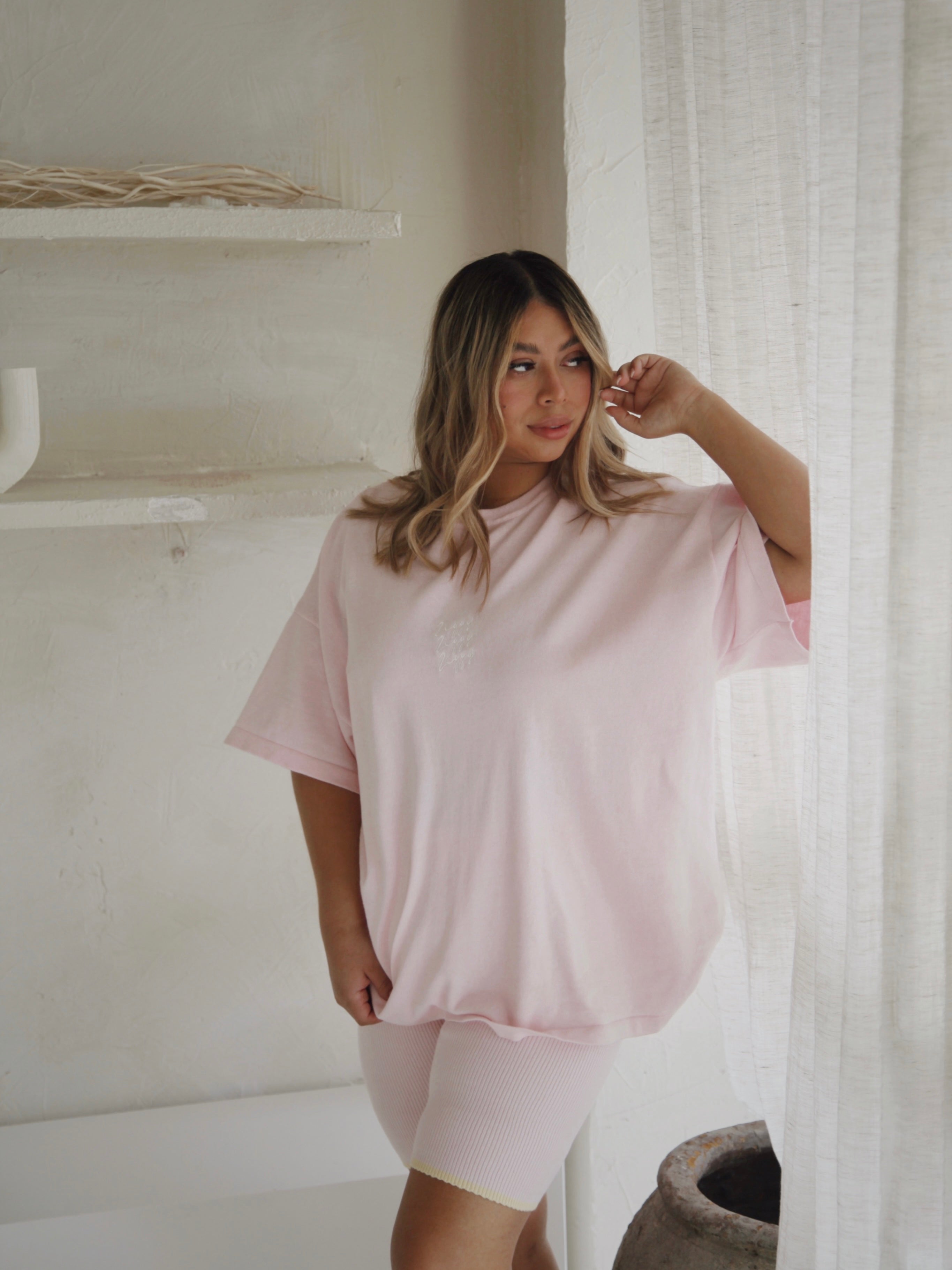 A woman wearing the Women's Signature Tee Freya from ZIGGY LOU, styled in an oversized fit and paired with shorts, stands by a window adorned with sheer curtains, gazing outside. The softly lit room has light-colored walls and minimal decor, including a simple shelf and a delicate vase in the corner.