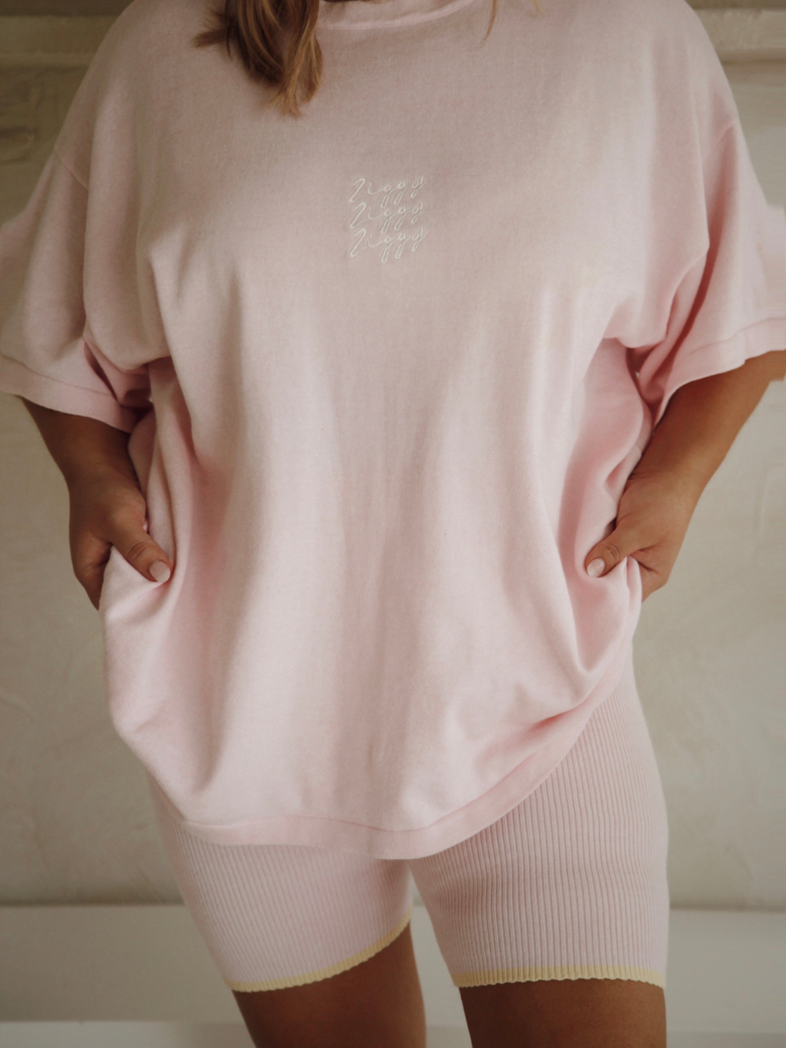 A person wearing a loose-fitting, light pink Women's Signature Tee Freya and matching ribbed shorts stands with hands in pockets. The oversized tee from ZIGGY LOU features the Ziggy embroidered logo on the chest in subtle white text. The background is softly blurred.