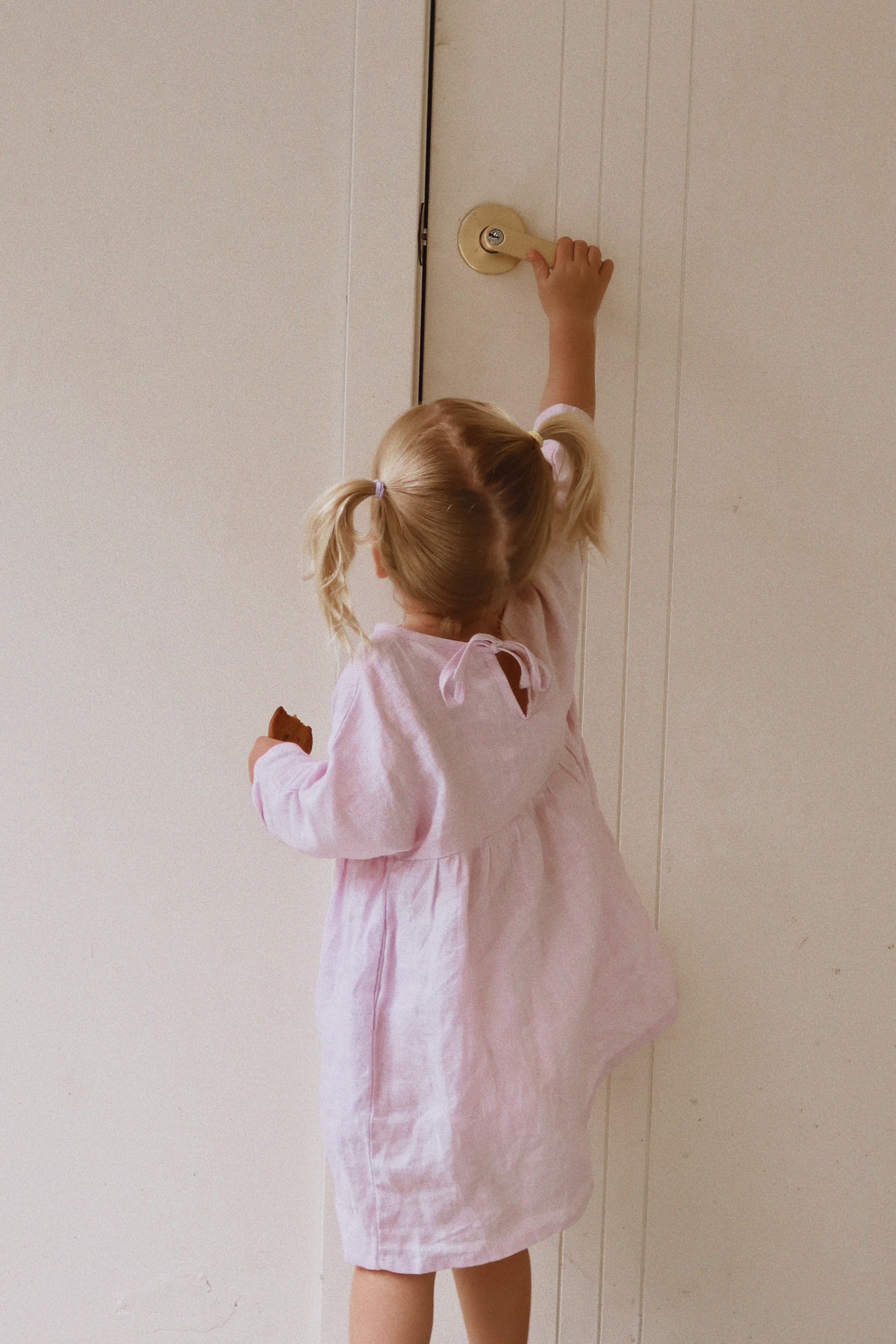 A small child with blonde pigtails, wearing the Posy Dress Lilac from MILKY DESIGNS in Size 5, reaches up to turn a doorknob on a white door. The knee-length dress flows gently as the child faces away from the camera.