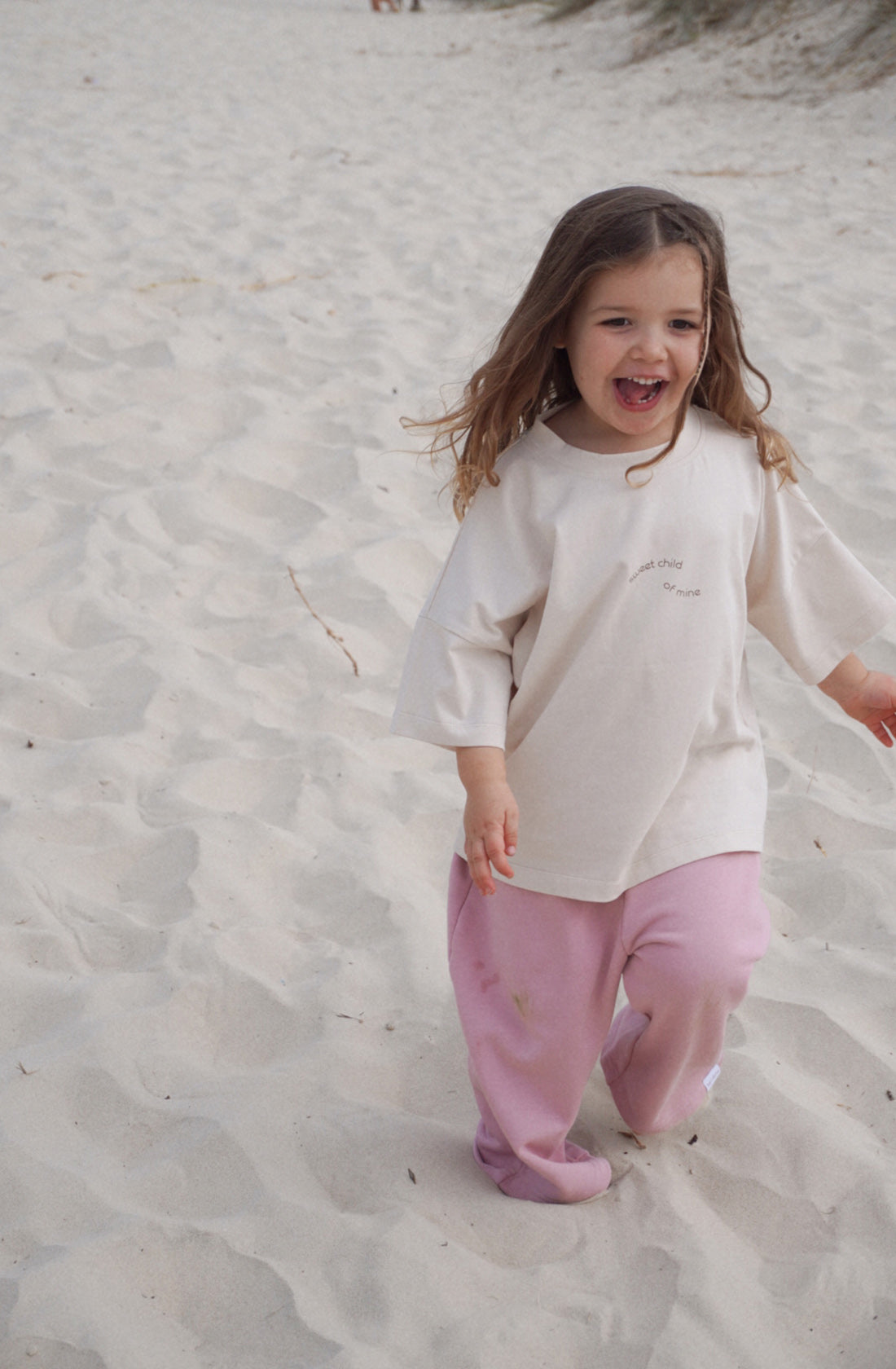 A joyful young child with long hair and a big smile walks on a sandy beach. The child is wearing the Sweet Child Tee by TWIN COLLECTIVE, an oversized beige t-shirt made from 100% cotton, paired with pink pants. With bare feet partially buried in the sand, the child looks carefree and happy.