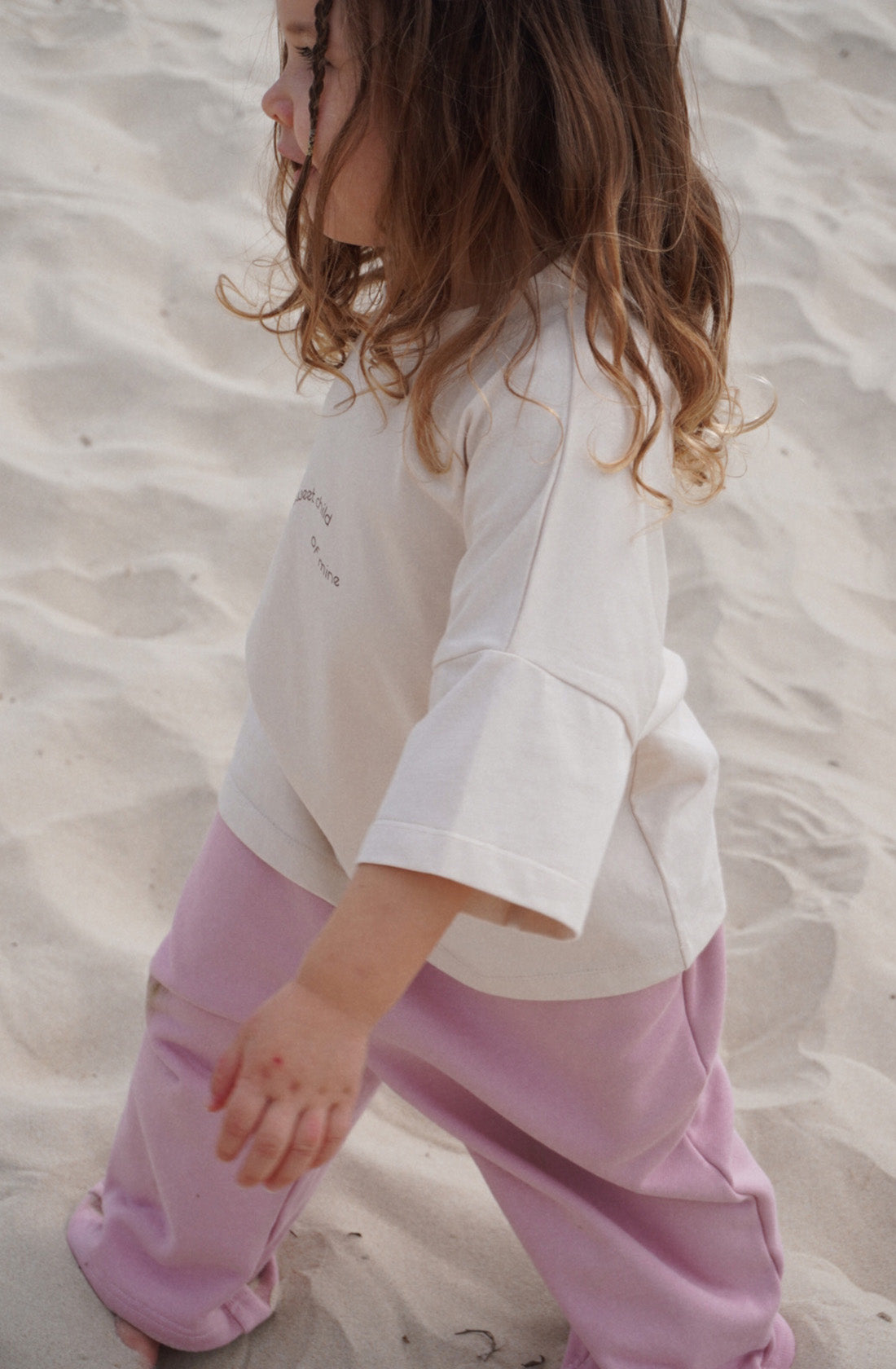 A young child with wavy, shoulder-length hair walks on sand in a white Sweet Child Tee from TWIN COLLECTIVE and pink pants. The 100% cotton shirt features an oversized fit, and the child appears to be focused on something in the direction they are walking.