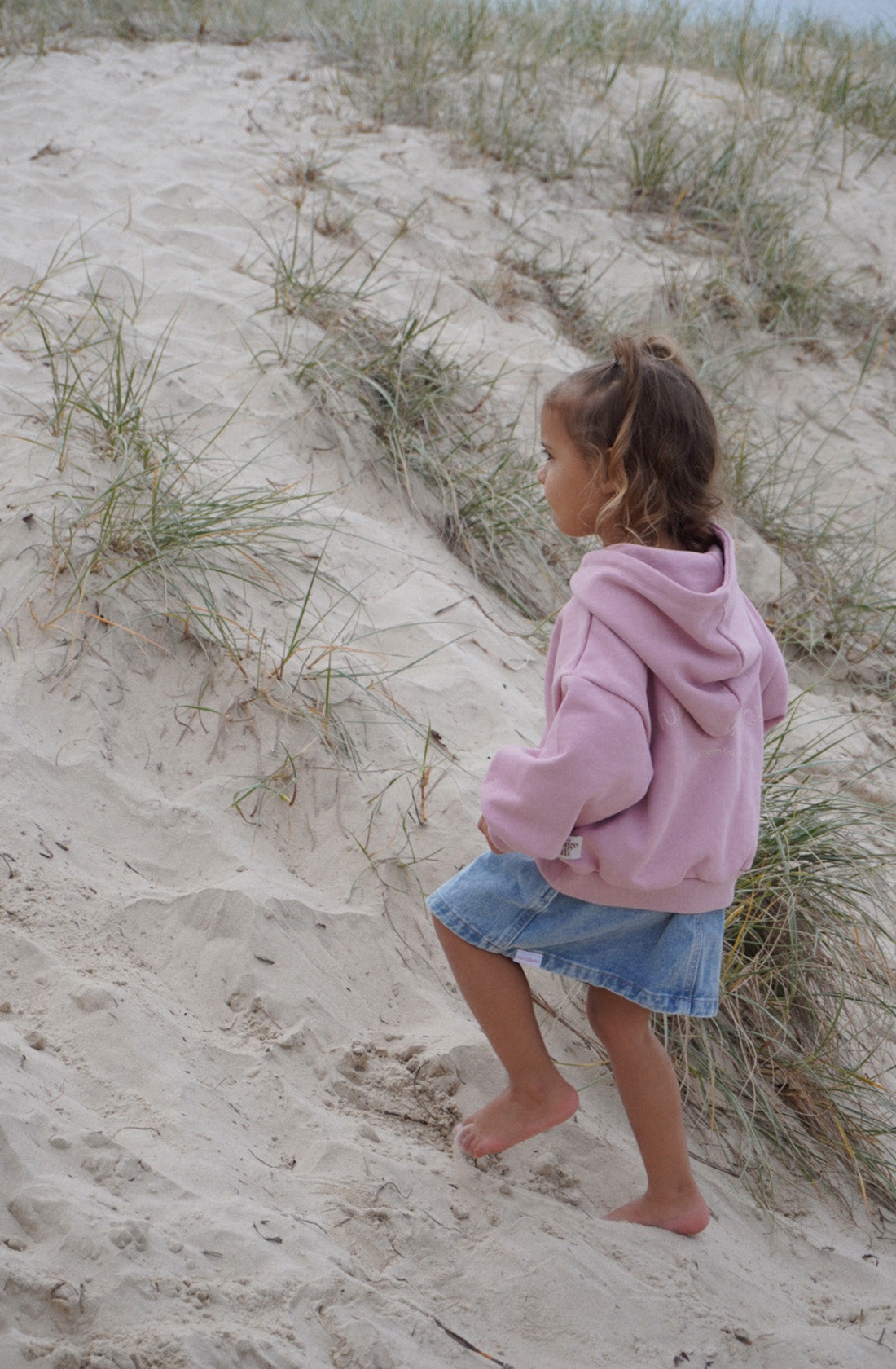 A young child with shoulder-length hair, wearing a TWIN COLLECTIVE Logo Hoodie Set Pink Haze and a denim skirt, is walking barefoot up a sandy dune with sparse grass. The child appears focused on the climb.