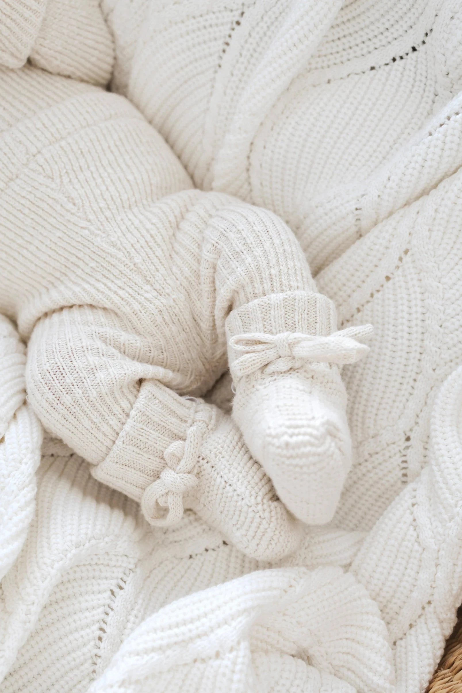 A close-up image of a baby's legs and feet, wearing knitted cream-colored pants and ZIGGY LOU's Booties Honey with bows, resting on a matching 100% textured cotton blanket.