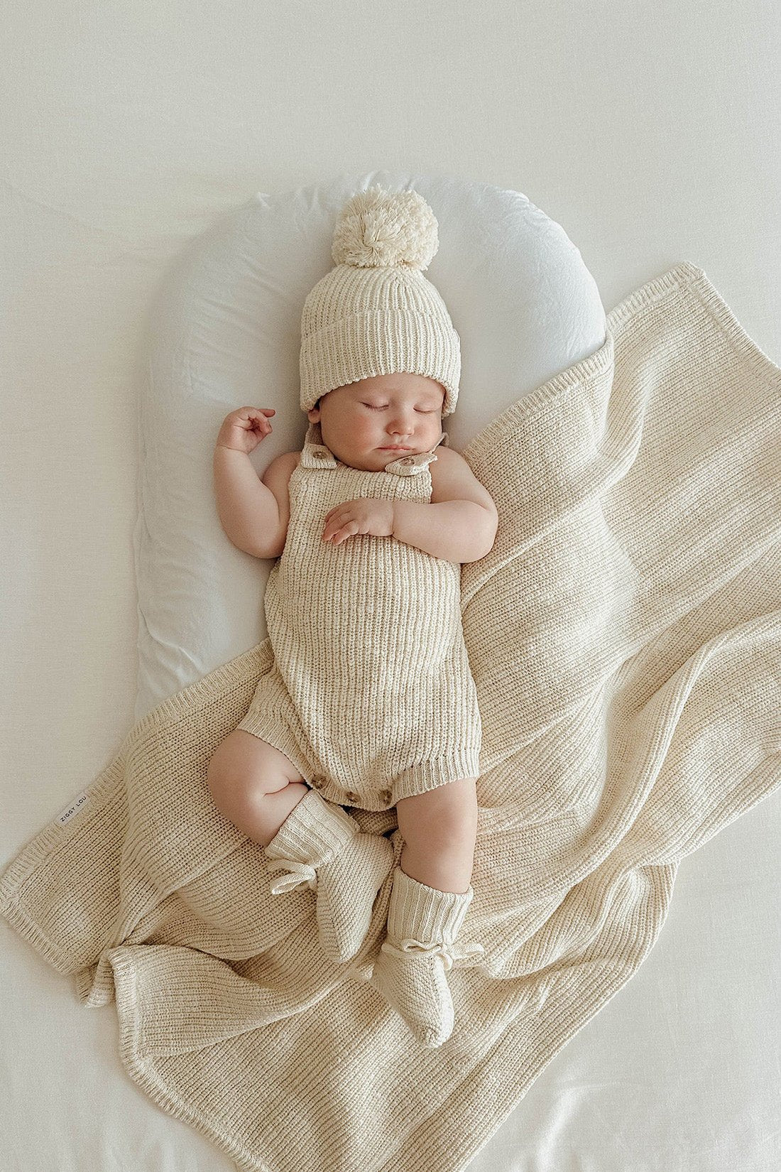 A small baby is sleeping peacefully on a white cushioned surface. The baby is dressed in a cozy, knitted beige outfit made of 100% textured cotton, with matching ZIGGY LOU heirloom booties and blanket. The hat has a pom-pom on top, and the baby is positioned on their back with one arm slightly raised.