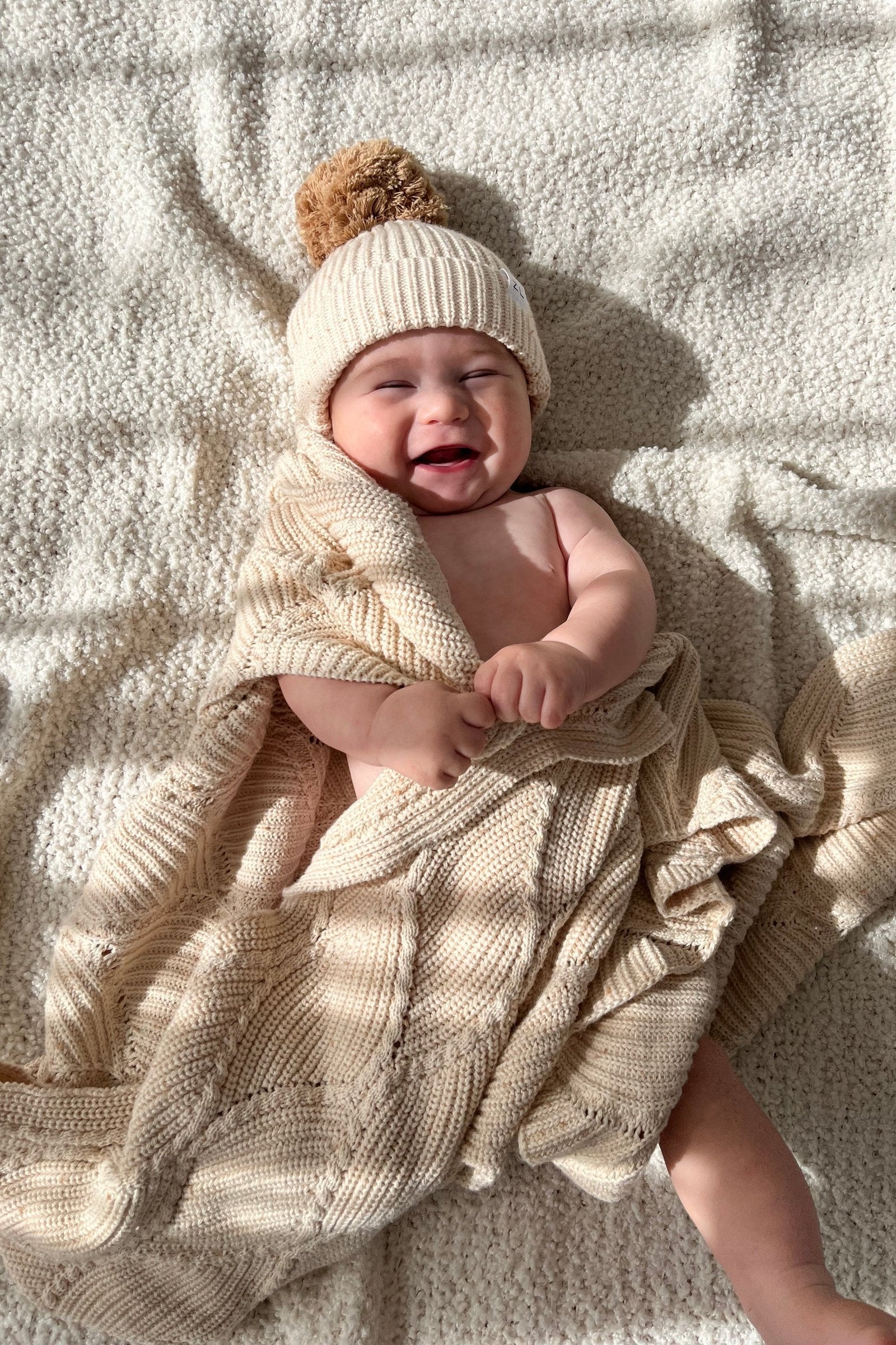 A baby wearing the ZIGGY LOU Beanie Biscotti Fleck, a chunky knit beanie with a pom-pom, is lying on a textured blanket. The baby is wrapped in a matching knit blanket and is smiling happily, showcasing an expression of joy. The soft lighting enhances the cozy and warm atmosphere reminiscent of biscotti fleck hues.