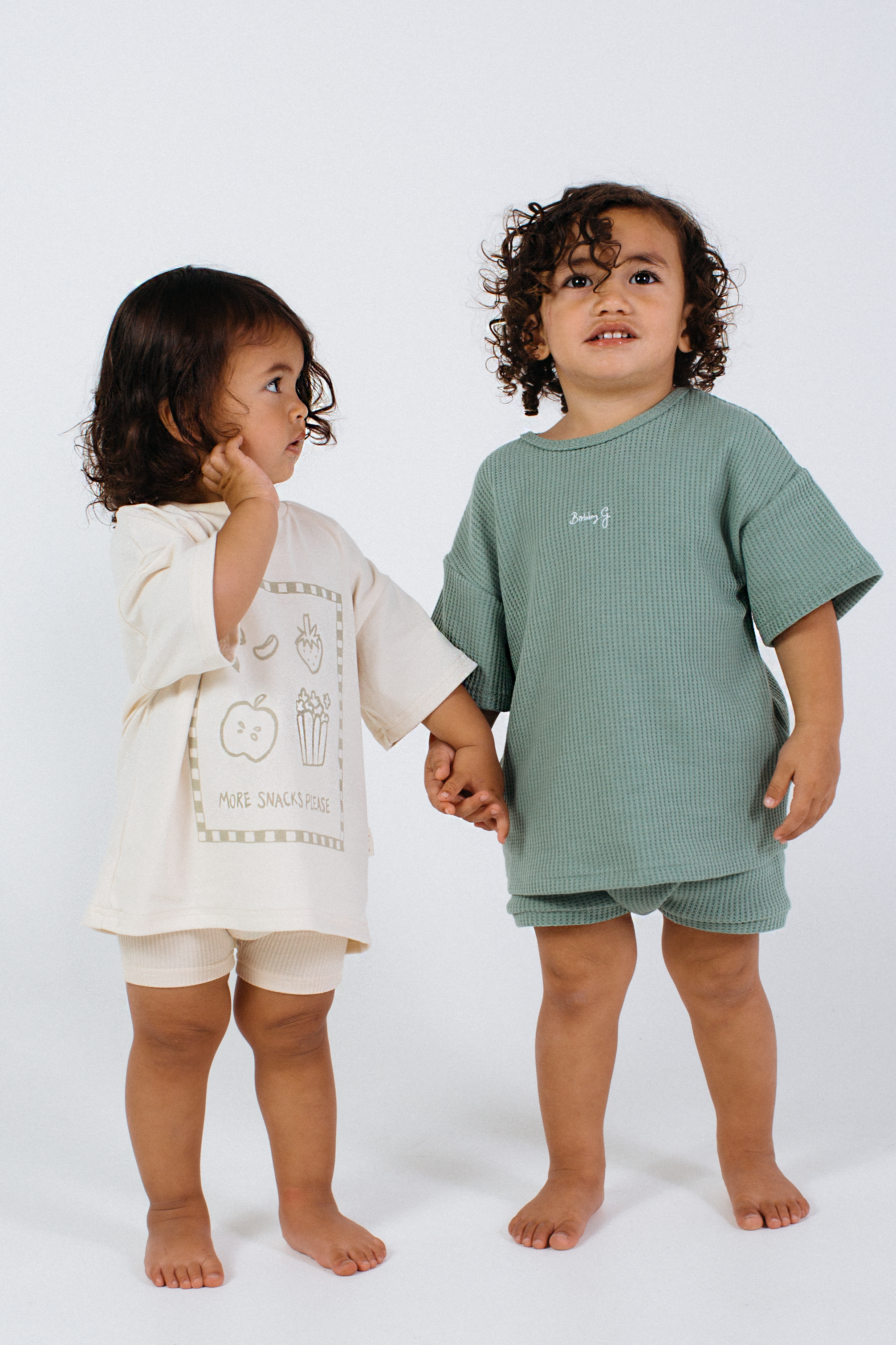 Two young children stand hand in hand on a white background. The child on the left sports a beige outfit with a graphic print, while the one on the right wears green Bike Shorts in Lemon Sherbet from BOBBY G BABY WEAR. Both have curly hair and seem to be looking in different directions, adding charm to their standout kids wardrobe.