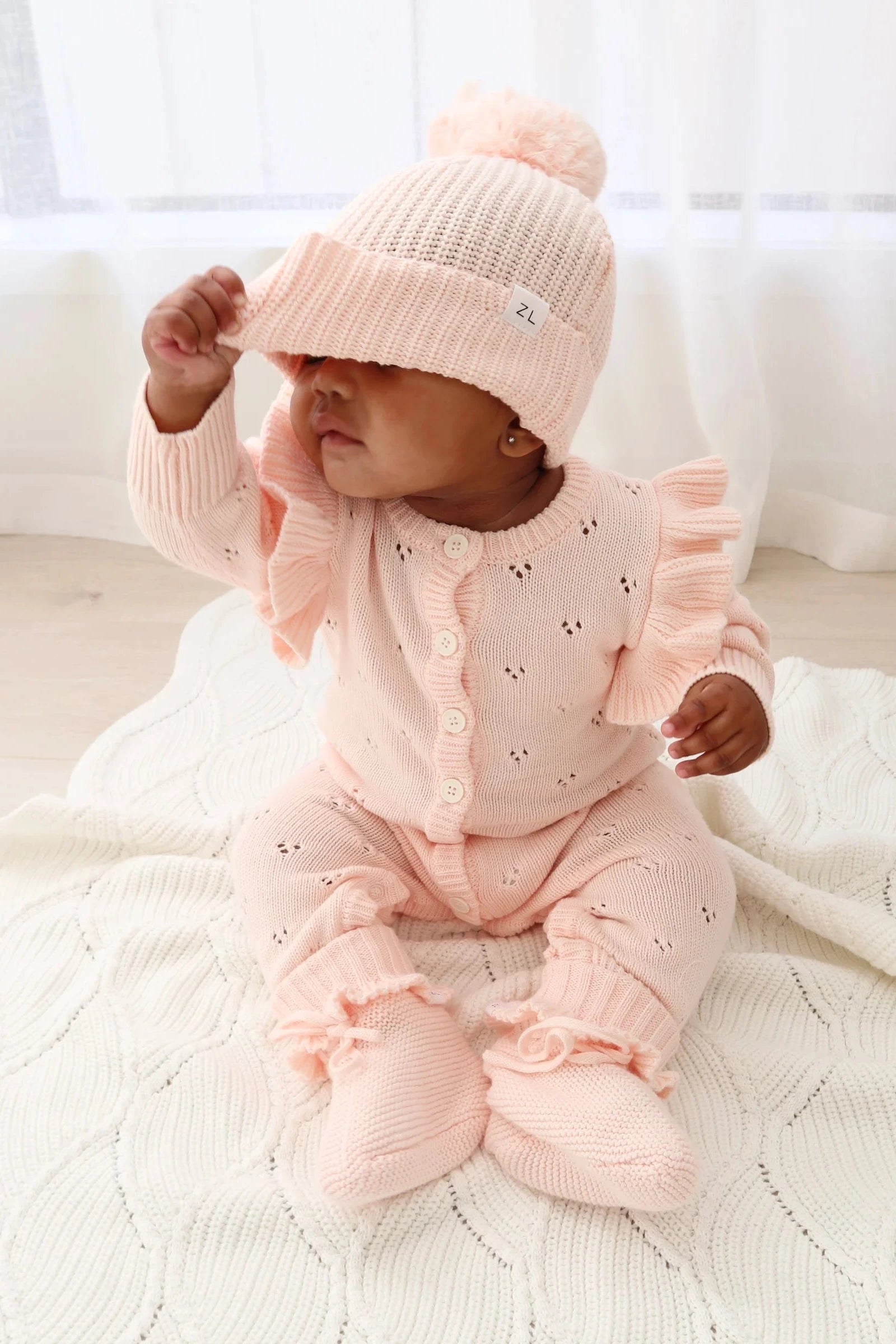 A baby sitting on a knitted blanket is dressed in a light pink outfit, complete with a matching ZIGGY LOU Beanie Primrose. The baby is lifting the edge of the beanie, partially covering their face. The room is softly lit with sheer white curtains in the background.