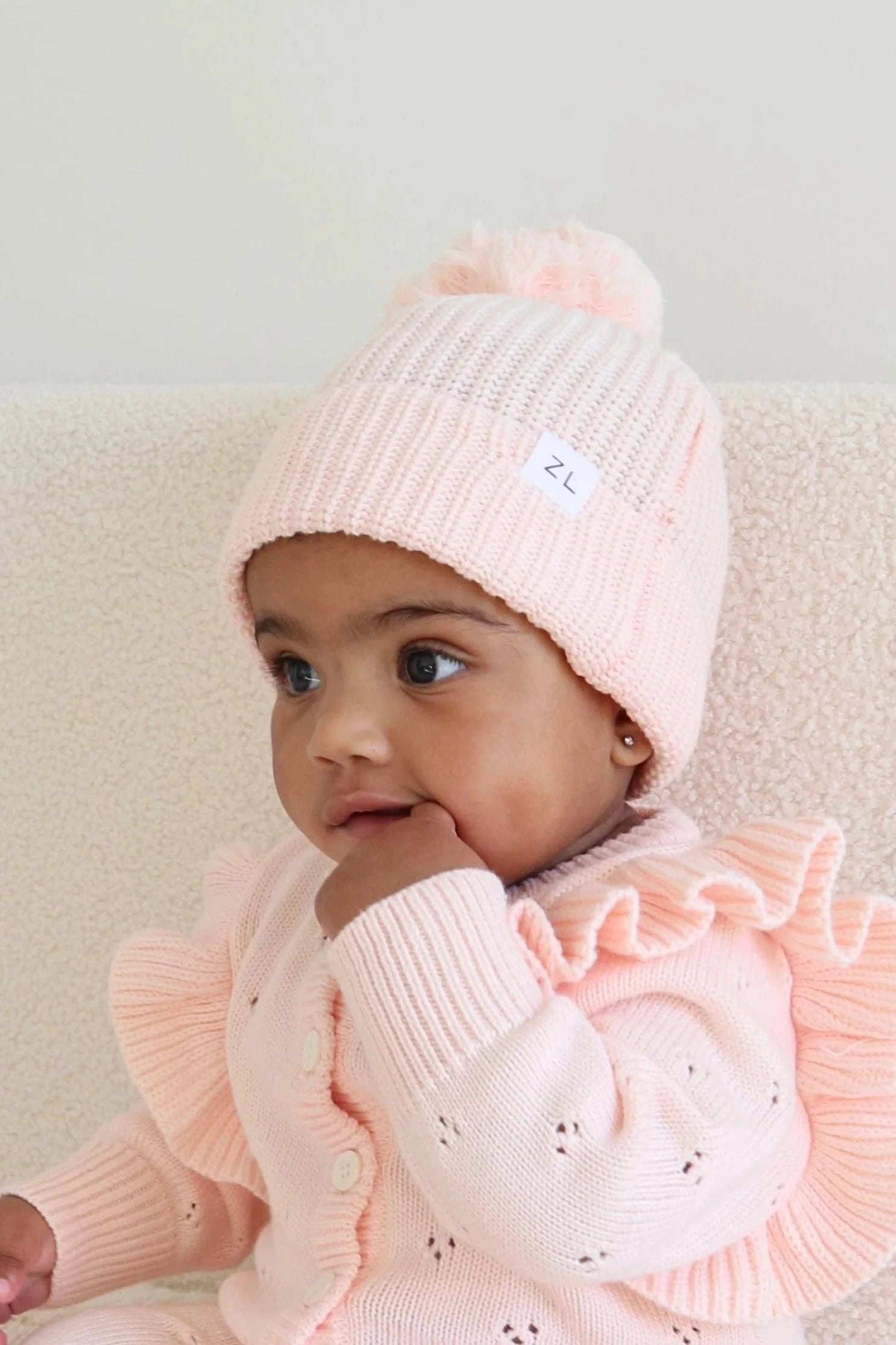 A baby dressed in a pink knit outfit with ruffled sleeves and wearing the Beanie Primrose chunky knit beanie from ZIGGY LOU, made from 100% cotton, sits against a light background. With a thoughtful expression and one hand raised to their mouth, the baby's beanie features a small label with the letters "ZL".