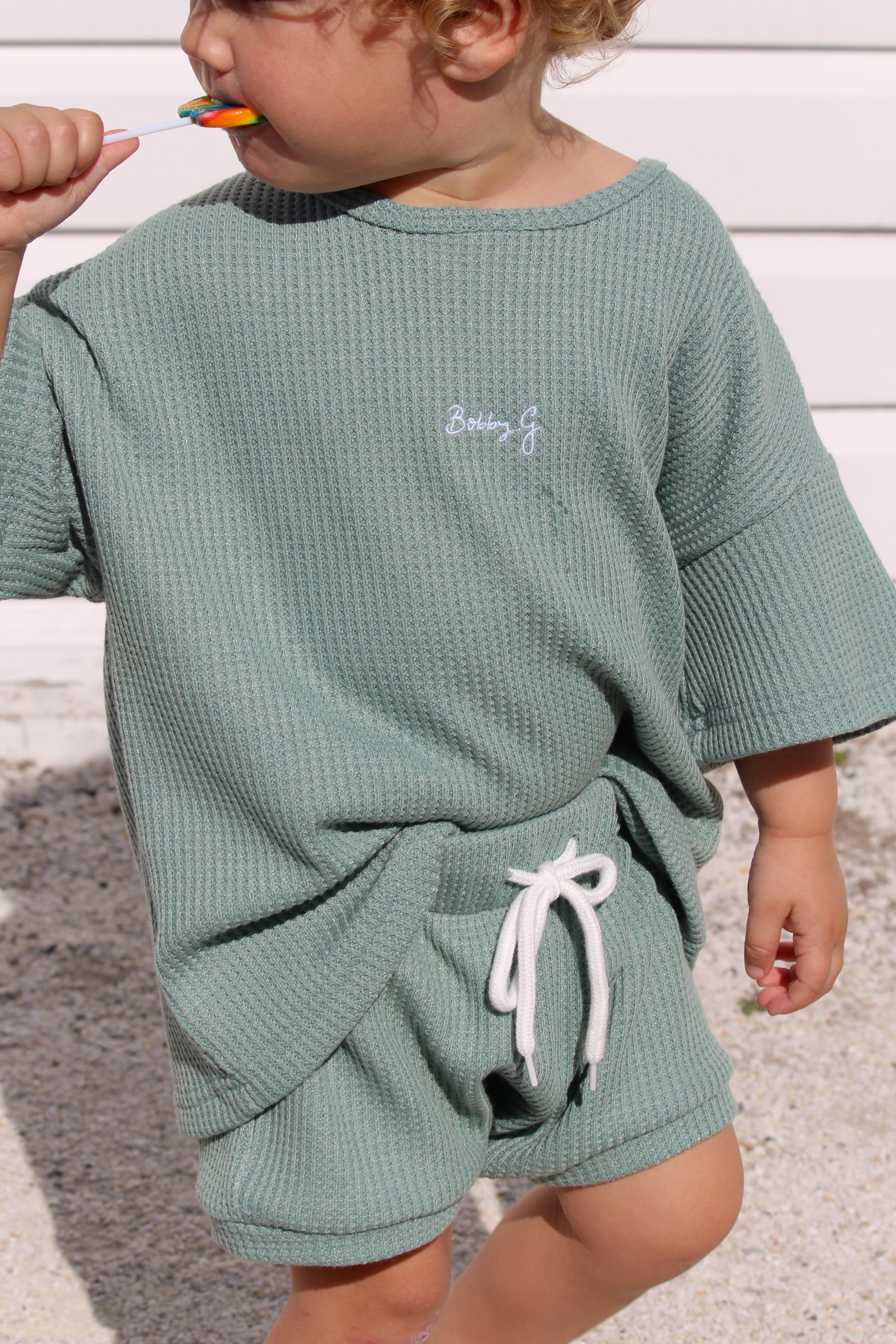 A toddler wearing oversized kiwi green Coby shorts from BOBBY G BABY WEAR paired with a waffle-knit t-shirt featuring an elastic waistband stands outdoors. The child holds a lollipop, with "Baby G" embroidered on the shirt's upper right side, enjoying all-day comfort in the adorable outfit.