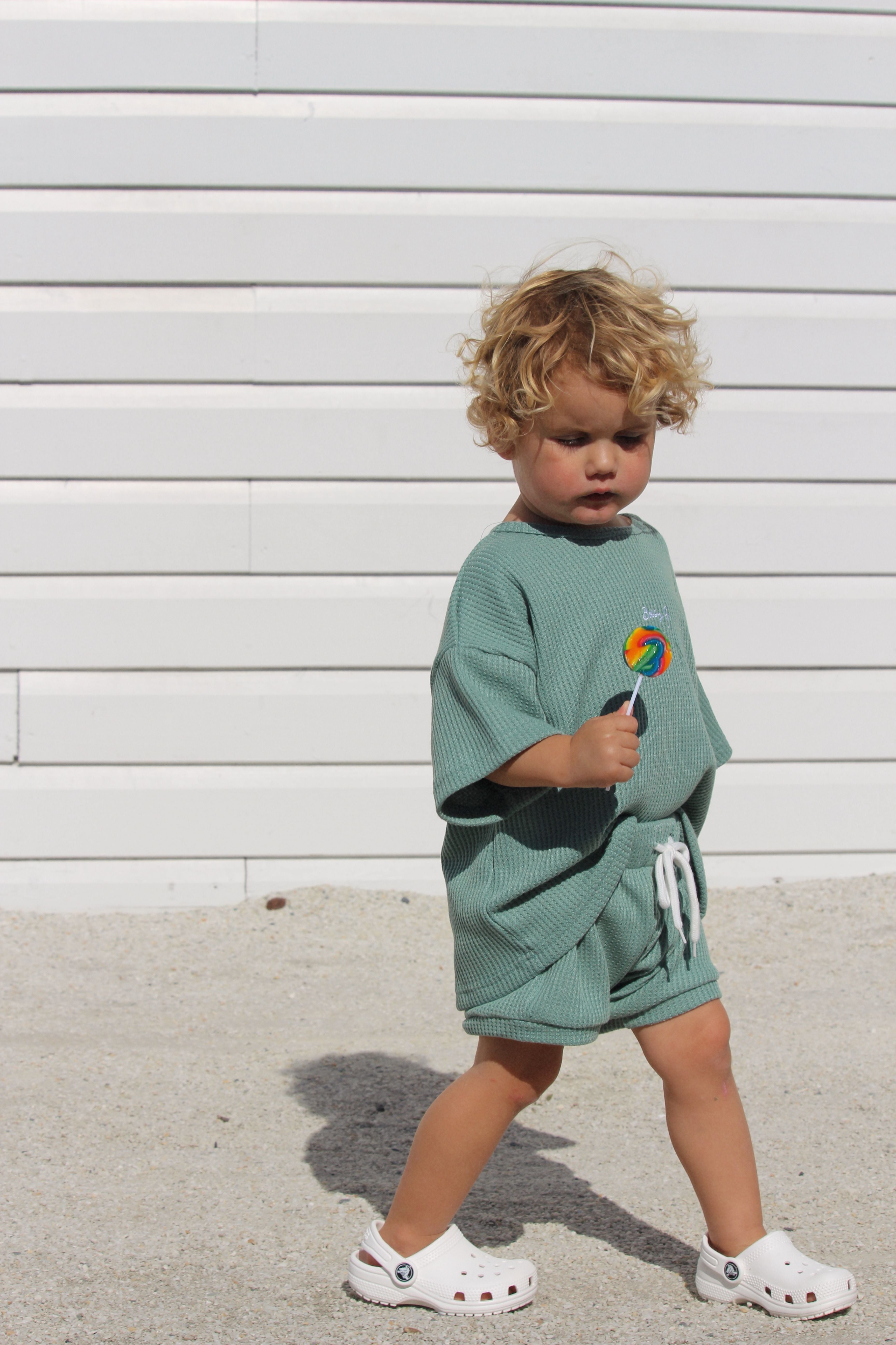 A young child with curly blond hair walks outdoors, wearing a light green outfit paired with Coby Shorts Kiwi by Bobby G Baby Wear. The shorts feature an elastic waistband for all-day comfort, and the child's look is completed with white sandals. The child is holding a lollipop against the backdrop of a white paneled wall and sandy ground.