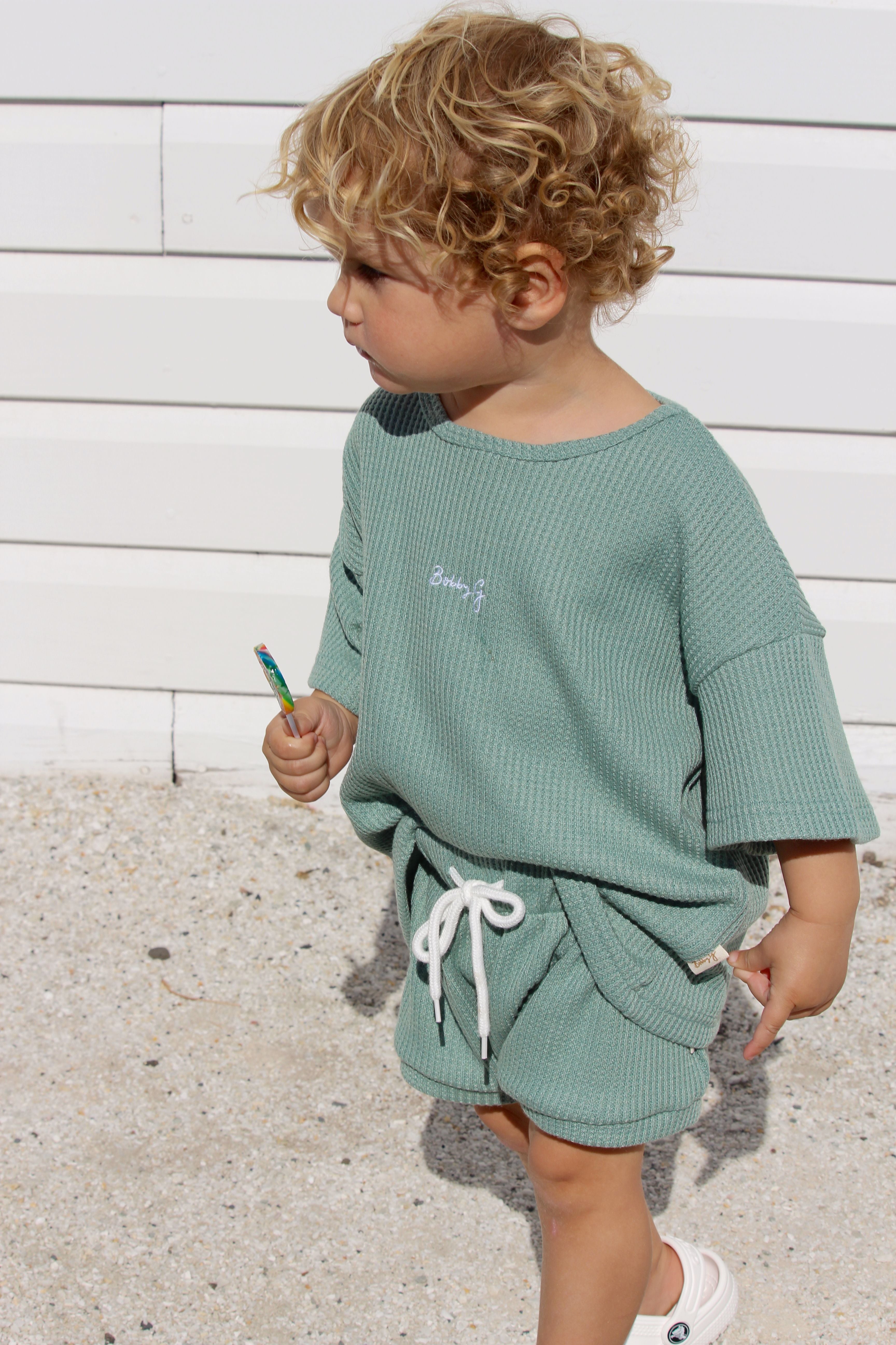 A young child with curly hair stands in front of a white wooden wall, wearing mint green Coby Shorts Kiwi from BOBBY G BABY WEAR, featuring an elastic waistband and a matching short-sleeved top. Holding a small toy in their hand, the child gazes to the side with a thoughtful expression, radiating all-day comfort.