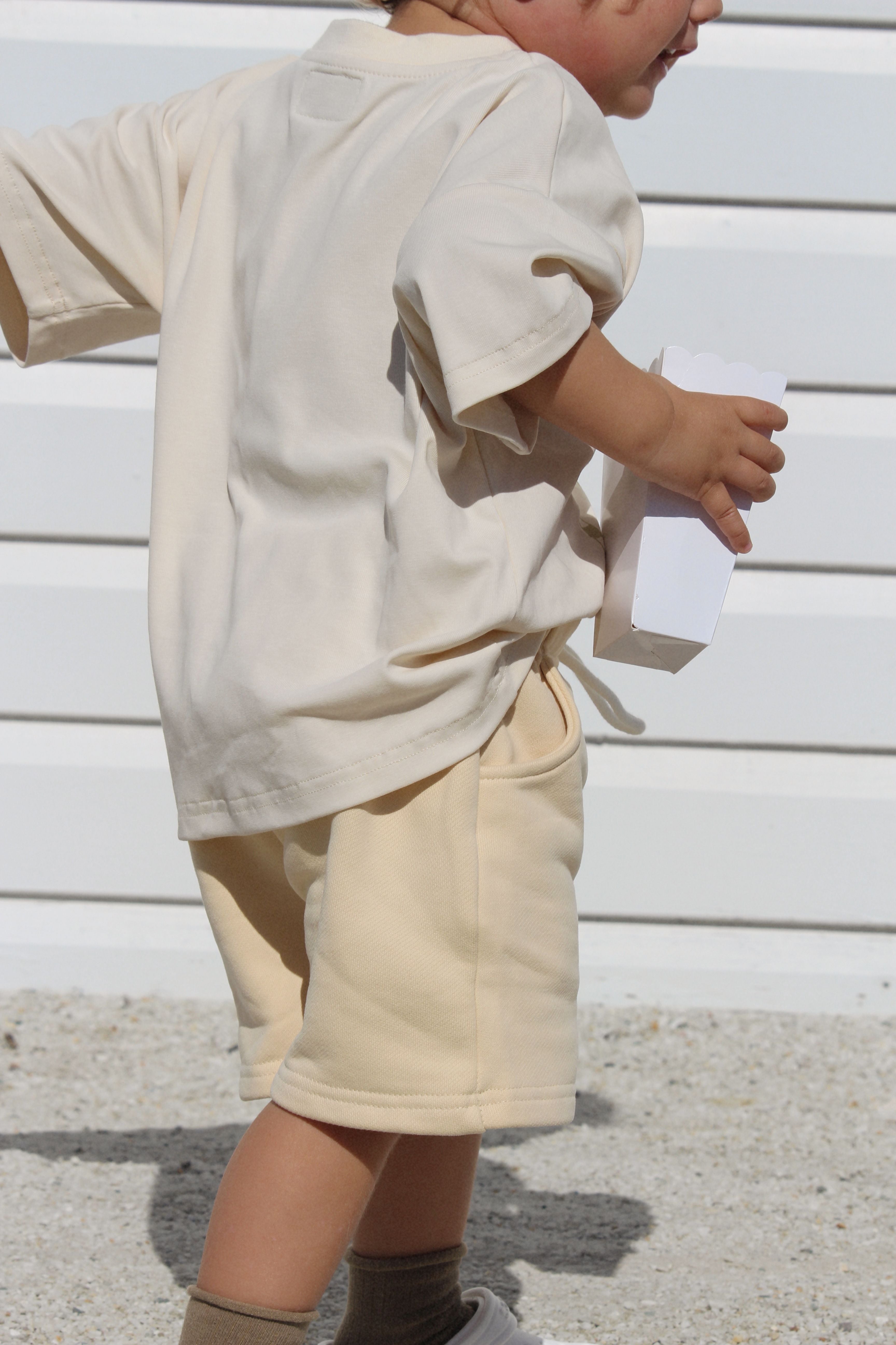 A young child wearing stylish and comfortable BOBBY G BABY WEAR's Frankie Shorts Popcorn, made from thick French terry fabric, is holding a paper object while standing on a gravel surface. The background features a white, horizontal panel wall, with the child’s face only partially visible.