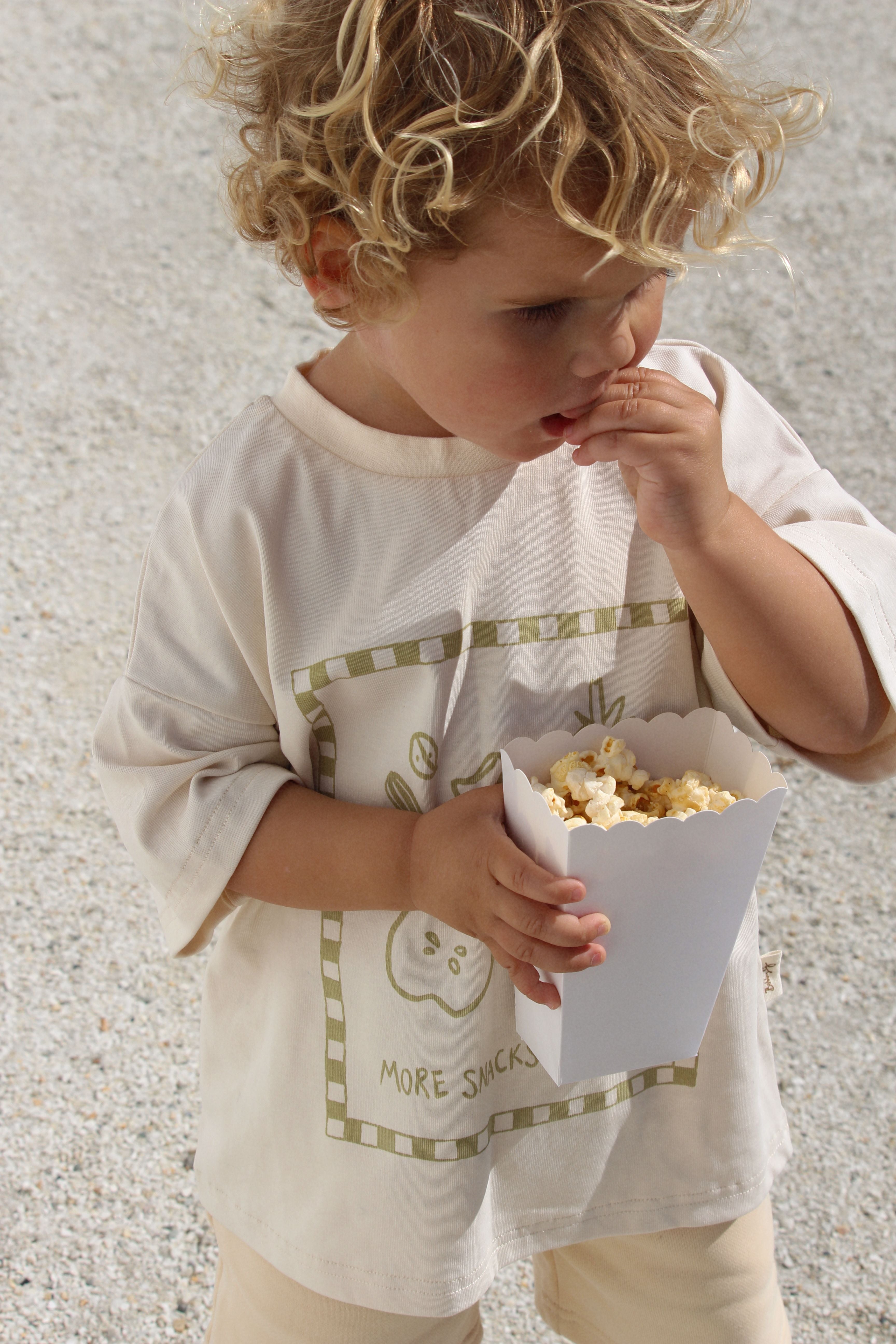 A child with curly blond hair is standing and eating popcorn from a white paper container. The child is wearing an oversized fit BOBBY G BABY WEAR 'More Snacks Please' Tee in Banana, made from organic cotton. The background appears to be an outdoor setting with a gravel surface.