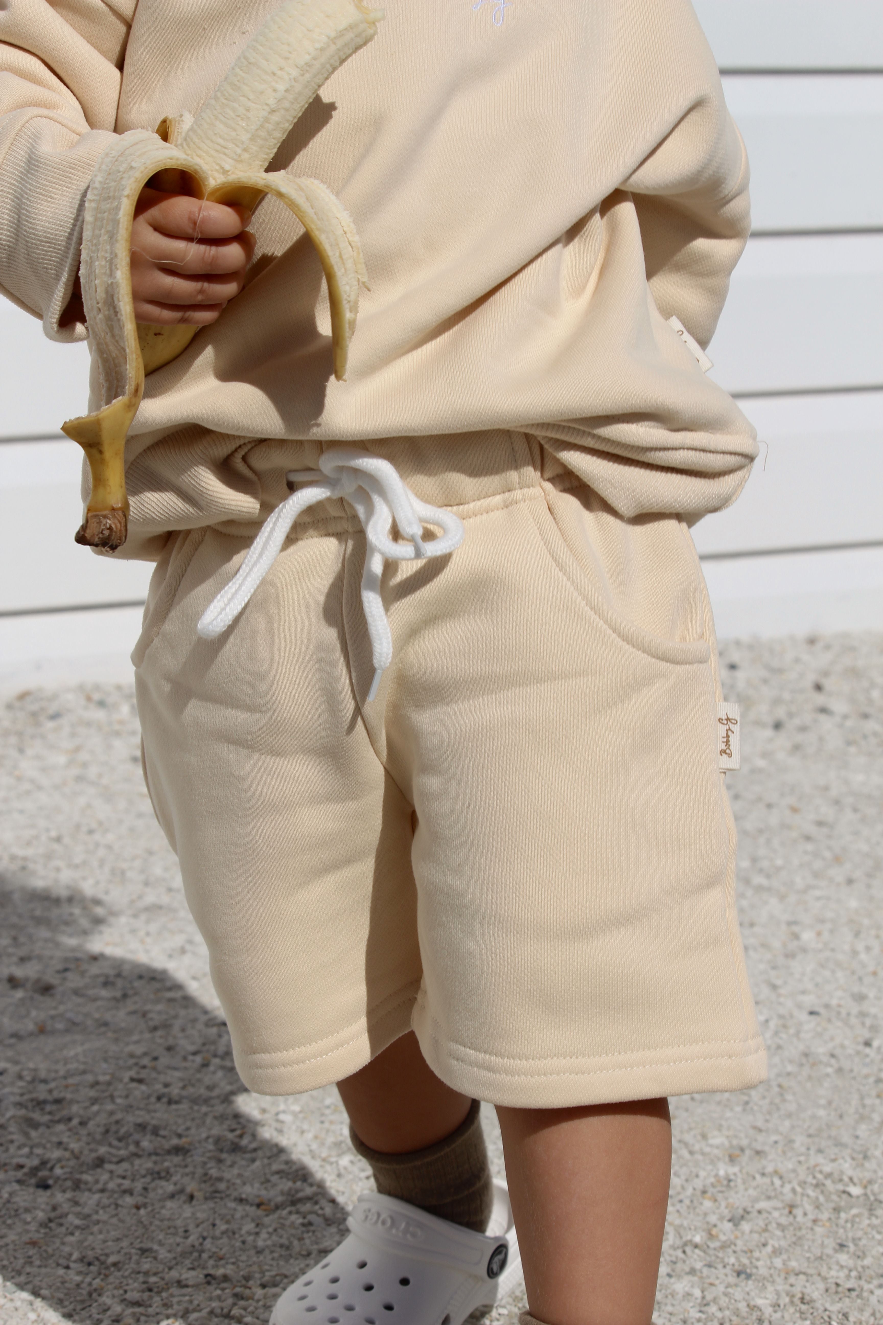 A child wearing a beige long-sleeve shirt and BOBBY G BABY WEAR's Frankie Shorts Popcorn holds a peeled banana. The child is standing on a light-colored surface, possibly sand, in front of a white wooden wall, and is also wearing white slip-on shoes.