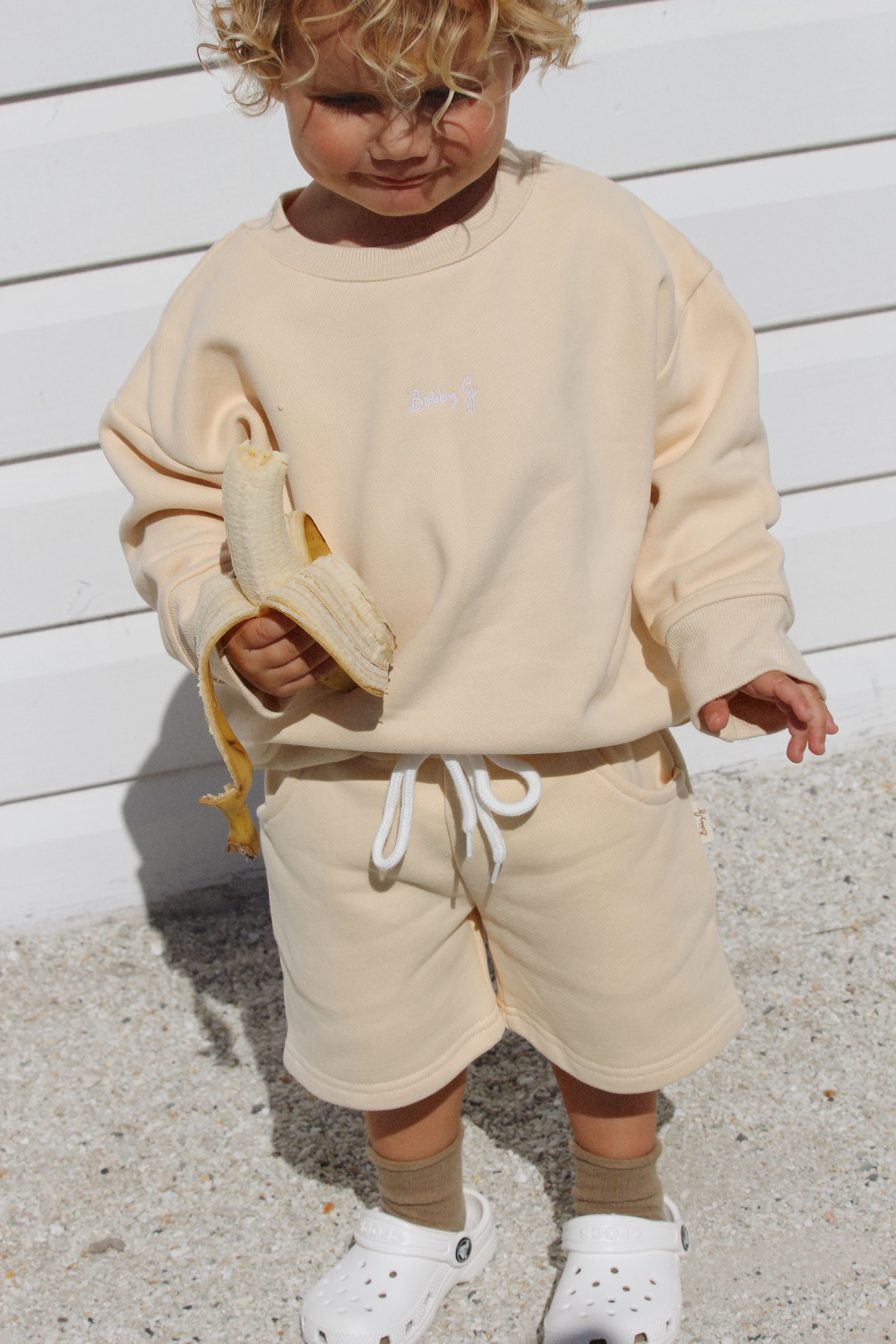 A young child with curly hair stands on a gravel surface, holding a peeled banana. They look stylish and comfortable in a light beige sweatshirt and matching Frankie Shorts Popcorn by BOBBY G BABY WEAR, crafted from soft French terry fabric. Their outfit is completed with white Crocs and light brown socks against a white paneled wall.