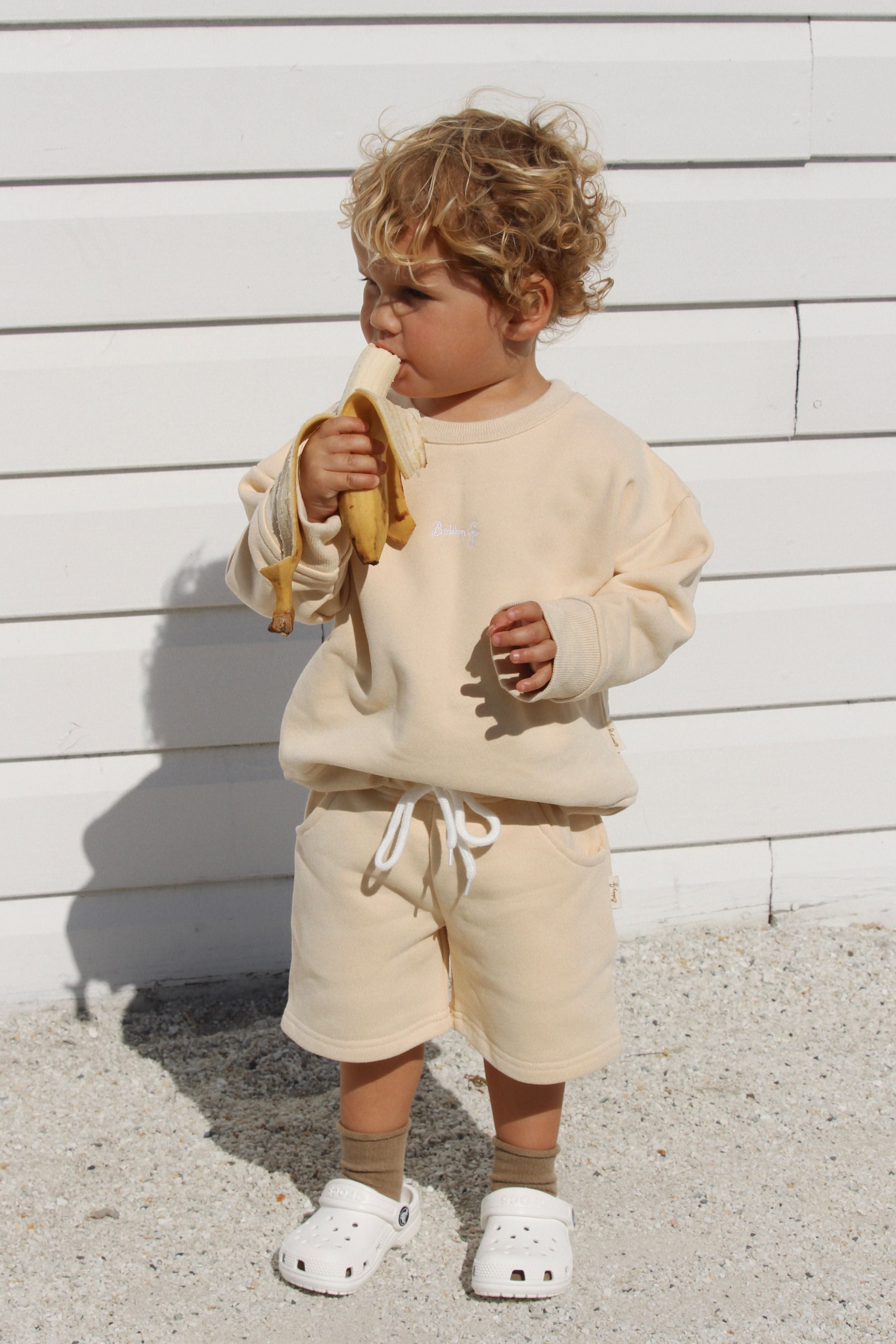 A curly-haired toddler stands on gravel, enjoying a banana while wearing the BOBBY G BABY WEAR Frankie Shorts Popcorn, complemented by a stylish beige French terry sweatshirt. They are photographed in front of a white wooden wall, completing the look with white clogs and socks.