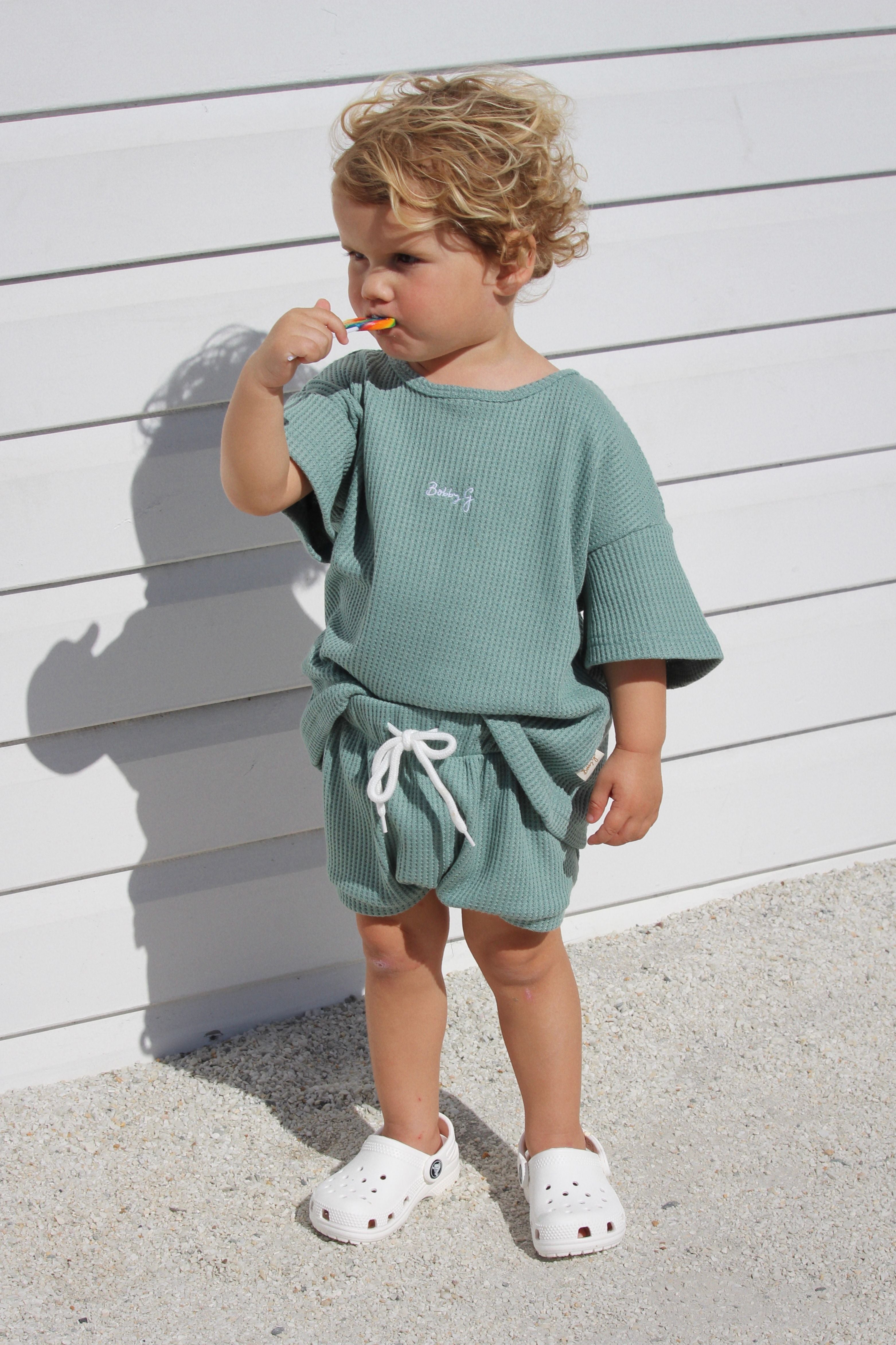 A young child with curly blond hair stands against a white wall, wearing BOBBY G BABY WEAR's Coby Shorts Kiwi with an elastic waistband and white shoes. This green outfit provides all-day comfort as the child sucks on a lollipop in their right hand, gazing thoughtfully to the side.