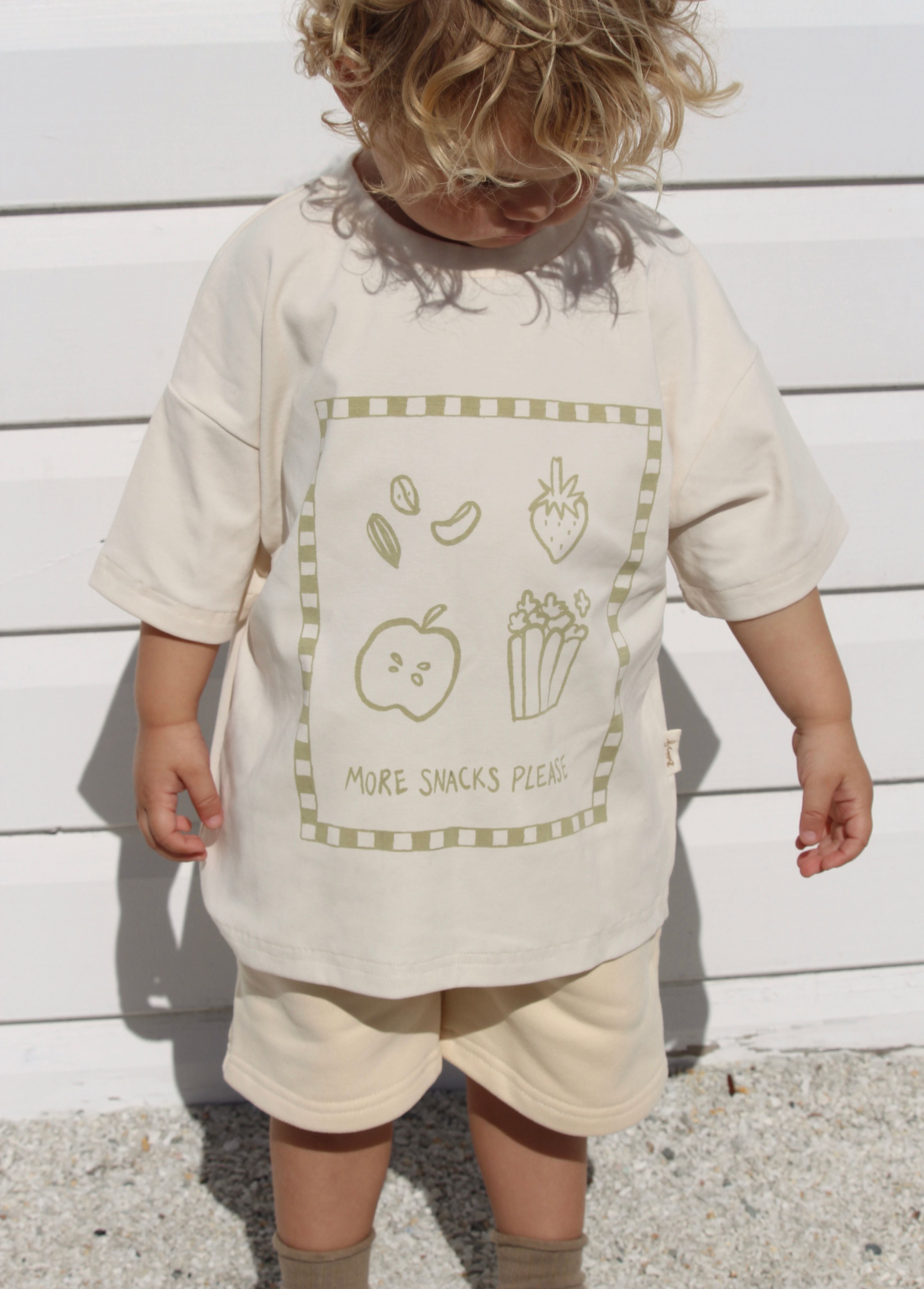 A young child with curly blonde hair stands in front of a white panel wall, wearing an oversized fit cream-colored T-shirt and matching shorts. The BOBBY G BABY WEAR 'More Snacks Please' Tee Banana features a simple drawing with snacks like an apple, strawberry, nuts, and popcorn, alongside the text "MORE SNACKS PLEASE.