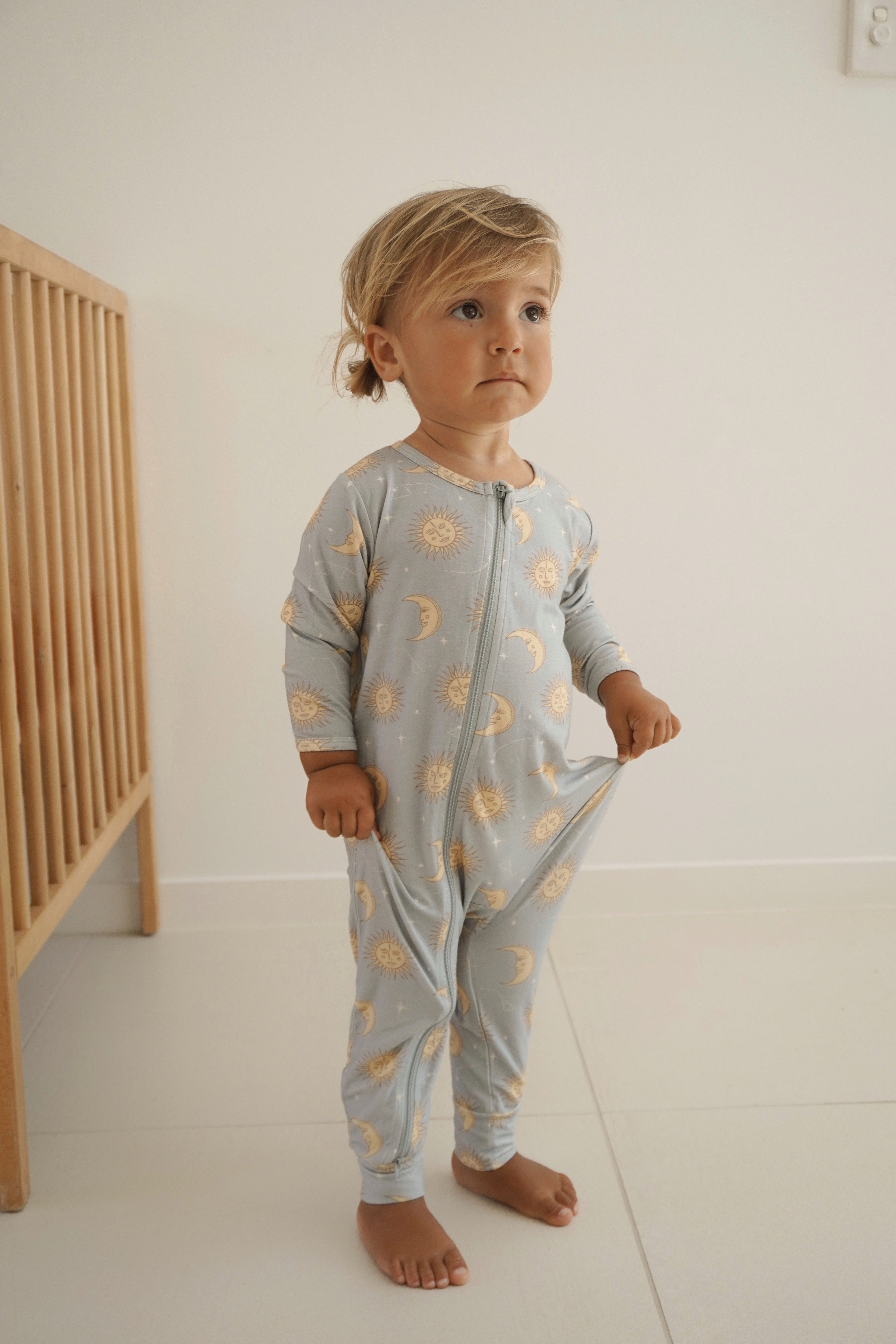 A young child stands in a "Celestial Zip Jumpsuit Dusty Blue" by BAM LOVES BOO, showcasing sun and moon patterns. With light brown hair and bare feet on a white tiled floor, the child clutches its sides. Nearby stands a wooden crib against the plain, light-colored background—a picture of serene innocence.