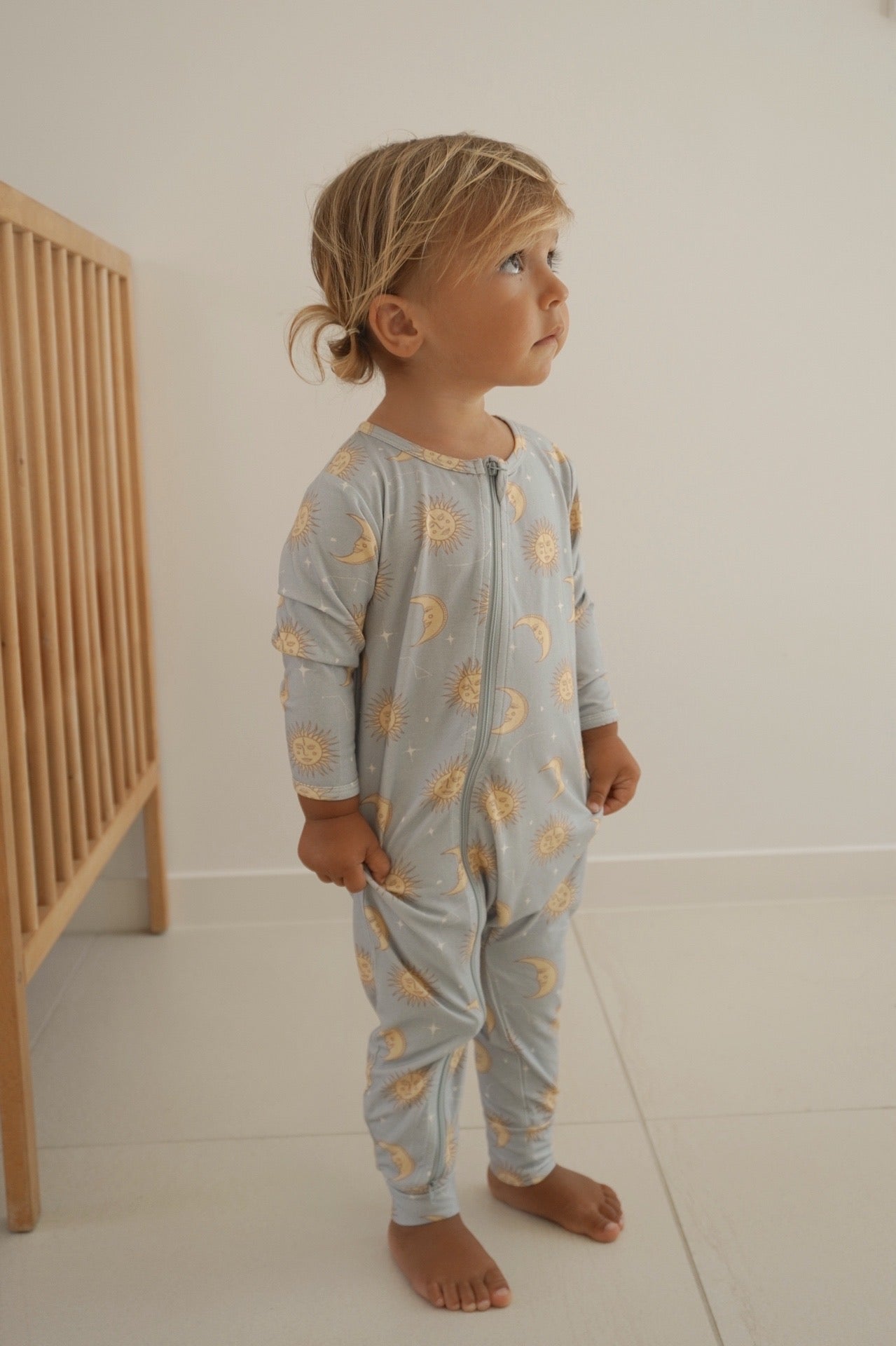 A toddler stands barefoot in a BAM LOVES BOO Celestial Zip Jumpsuit Dusty Blue, adorned with a celestial print, gazing upward. The child is beside a wooden crib against a light-colored wall, creating a soft and cozy atmosphere ideal for dreams and discoveries.