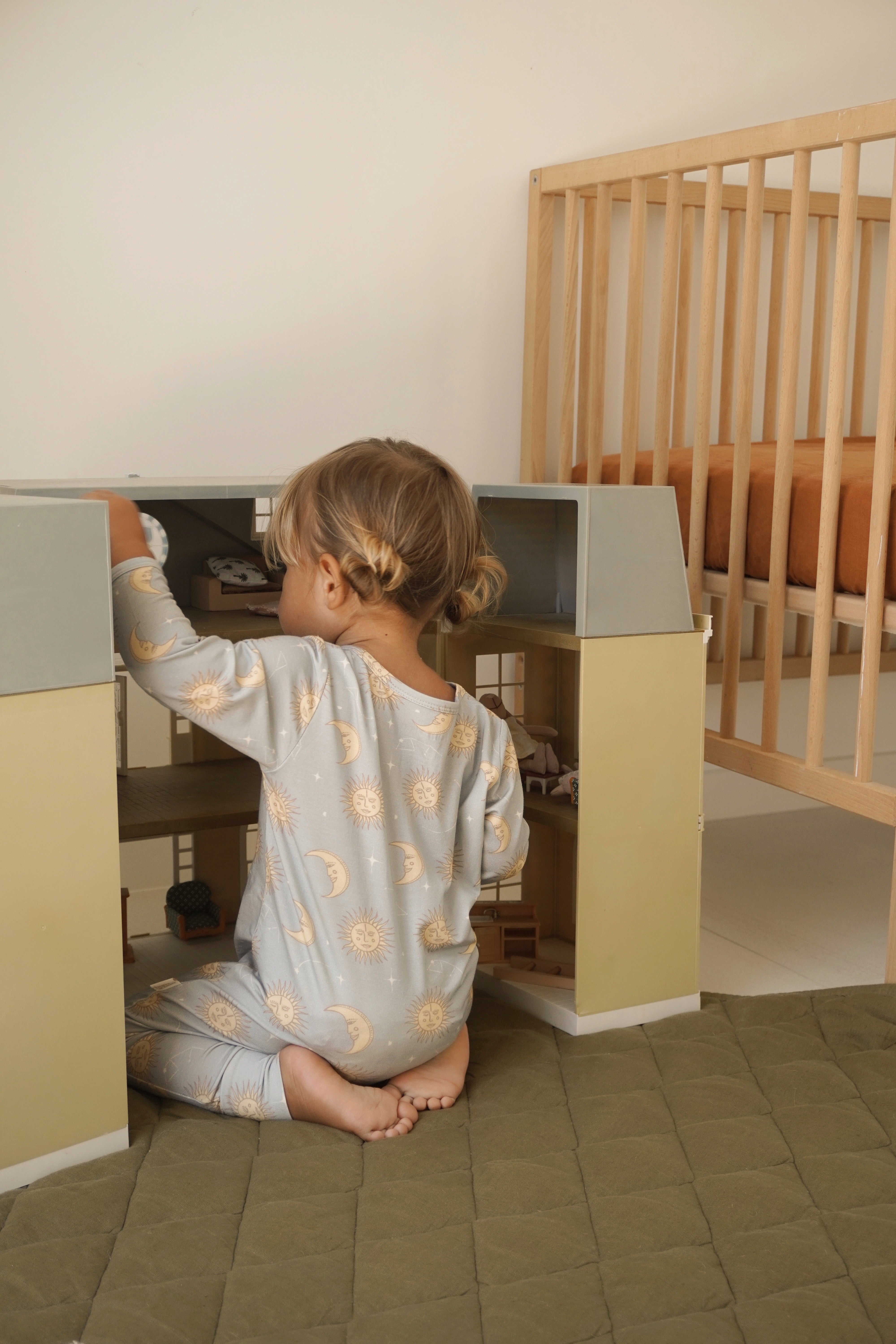 A young child with short hair, dressed in the BAM LOVES BOO Celestial Zip Jumpsuit in Dusty Blue, plays with a dollhouse on the floor. Beside the child is a wooden crib featuring an orange mattress. The environment is neutral and minimalistic.