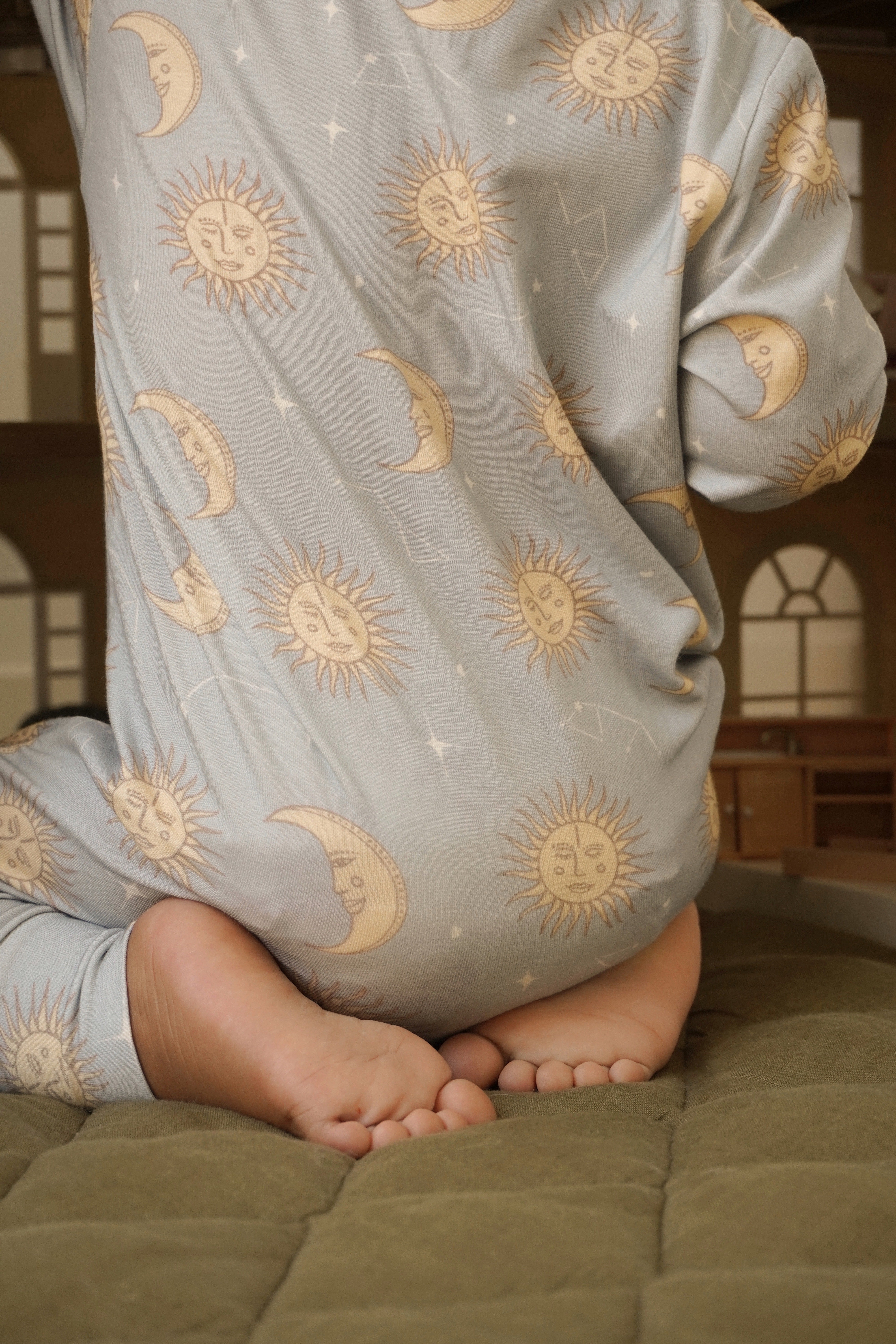 A child in a BAM LOVES BOO "Celestial Zip Jumpsuit Dusty Blue," featuring celestial prints of suns and crescent moons, sits on a quilted surface, facing away. The room in the background has soft lighting and arched windows.