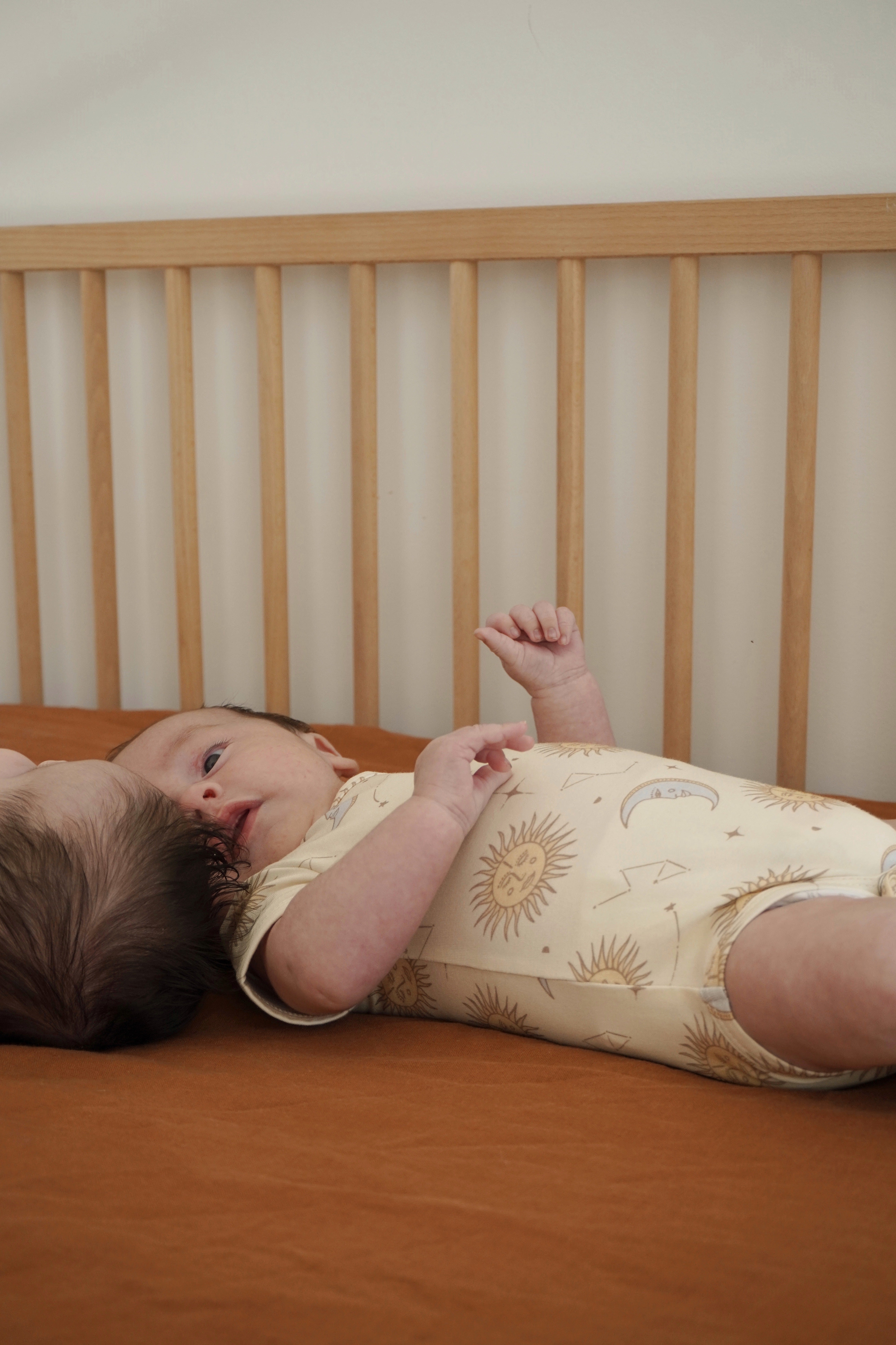 A baby rests peacefully on a brown bed, dressed in the "Celestial Short Sleeved Onesie Cream" from BAM LOVES BOO, featuring a celestial print made from bamboo organic cotton. The wooden slats of the crib can be seen in the background. The baby's expression is calm and relaxed.