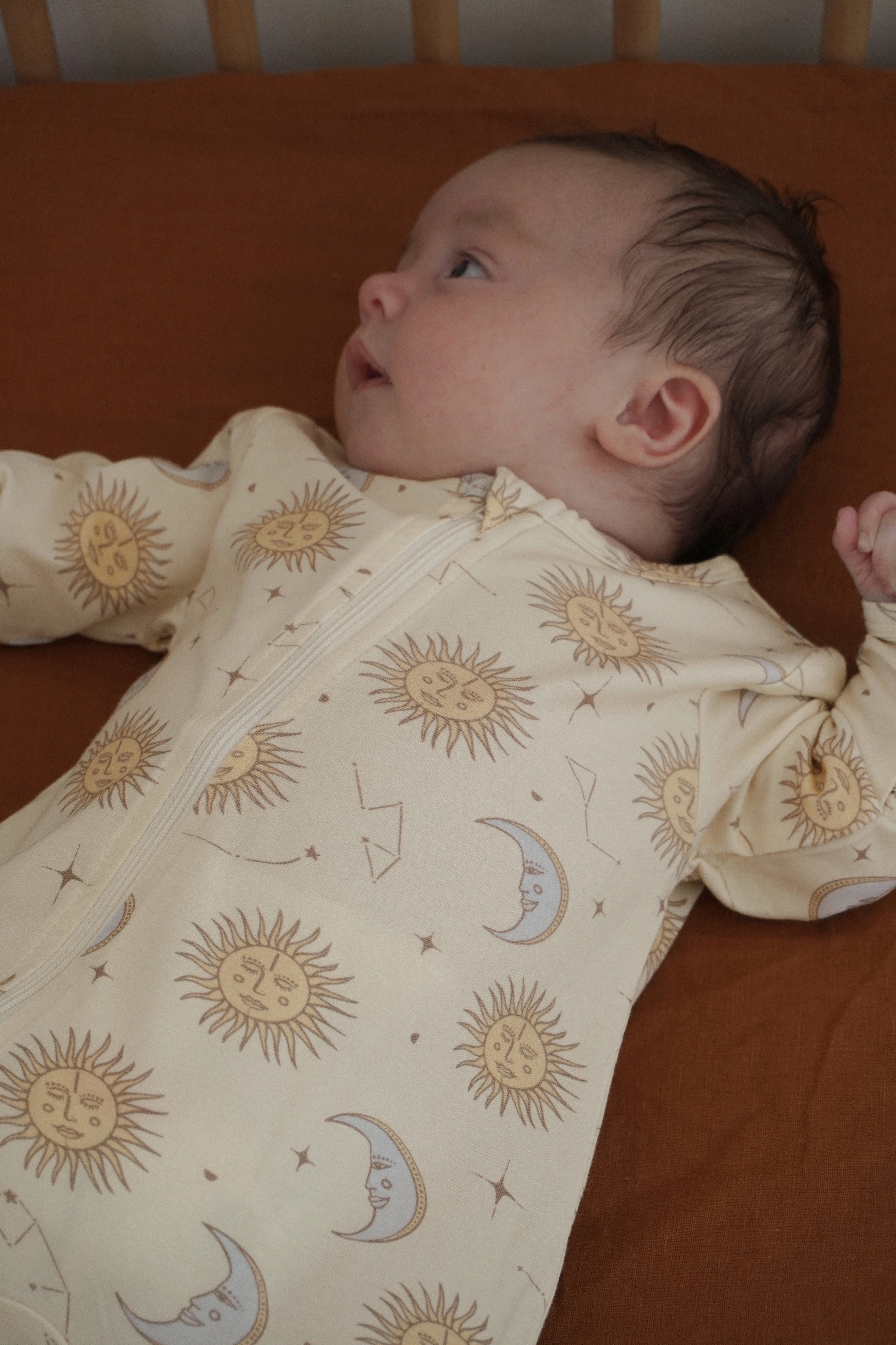 A baby peacefully rests on a brown surface, dressed in BAM LOVES BOO's Celestial Zip Jumpsuit Cream (Size 12-18M), featuring suns, moons, and stars. With their left hand raised slightly, the baby gazes upward in wonder.