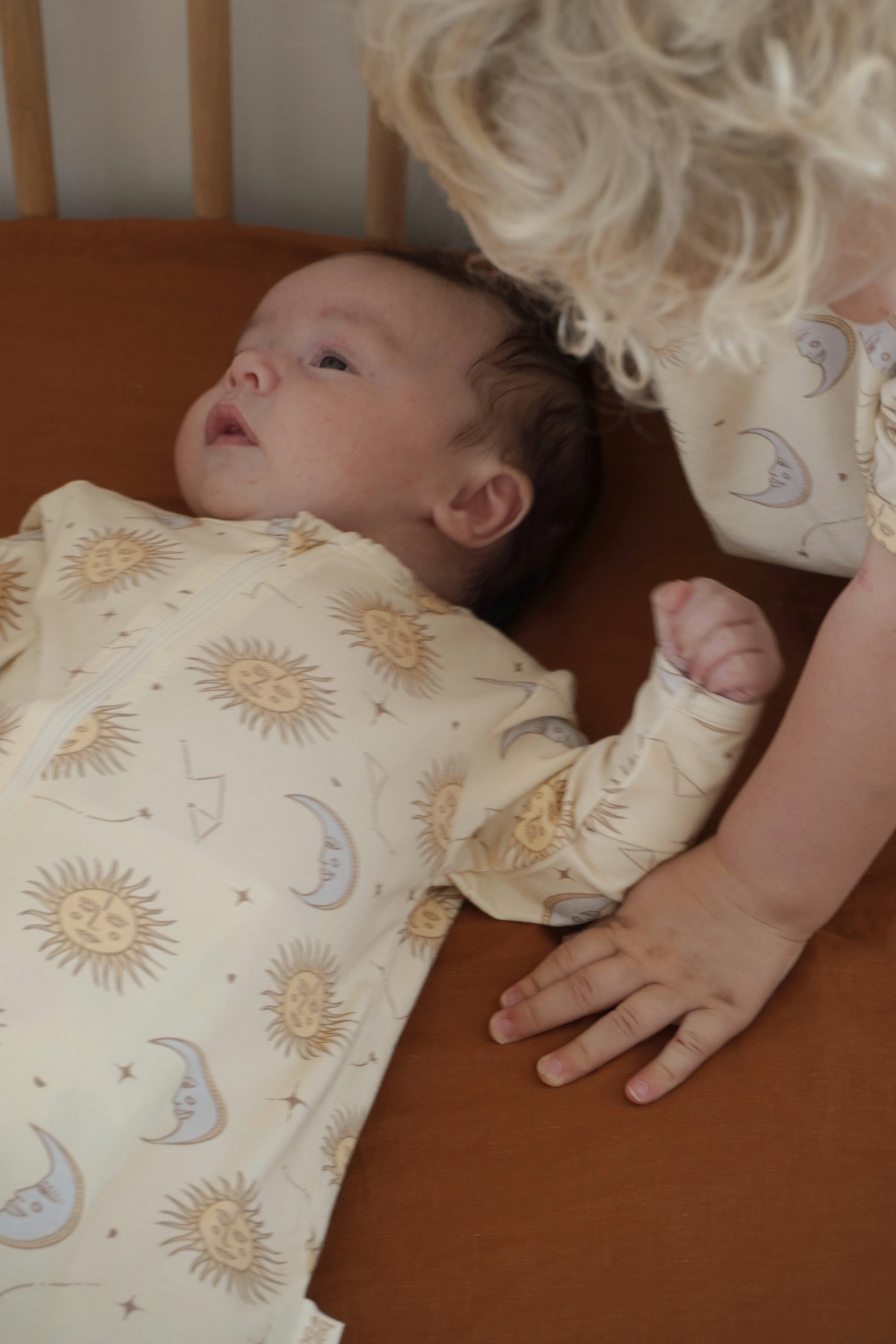 A baby lies on a brown surface wearing the Celestial Zip Jumpsuit Cream by BAM LOVES BOO, crafted from soft bamboo organic cotton and featuring a celestial print. A toddler with curly blonde hair, dressed in the same jumpsuit with a convenient two-way zipper, gently rests their hand on the baby's arm while gazing at them closely.