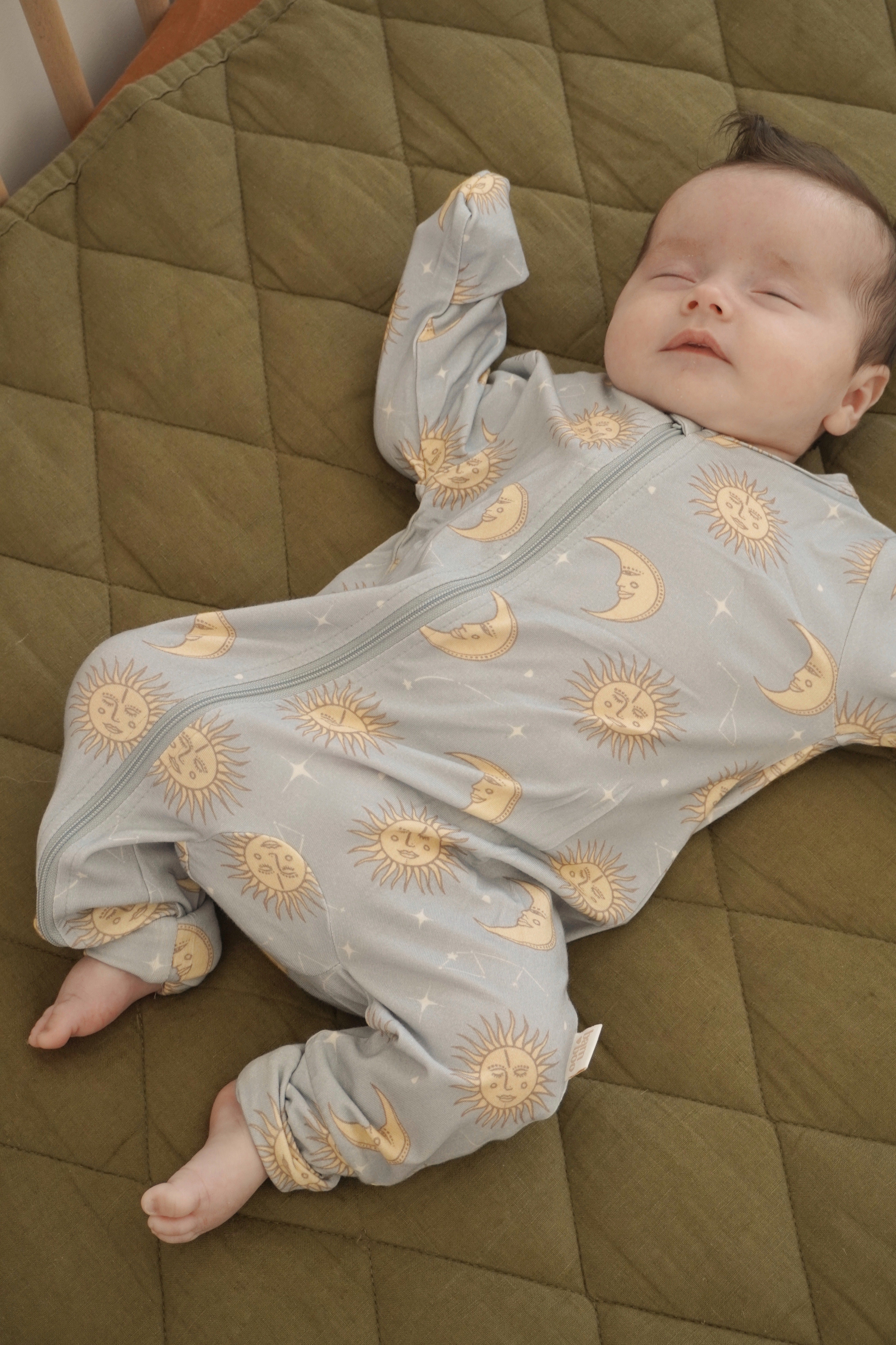 A baby sleeps peacefully on a quilted green blanket, wearing the Celestial Zip Jumpsuit in Dusty Blue from BAM LOVES BOO, adorned with a celestial print of suns and moons. With one arm raised above their head, the baby's gentle expression adds to the serene scene.