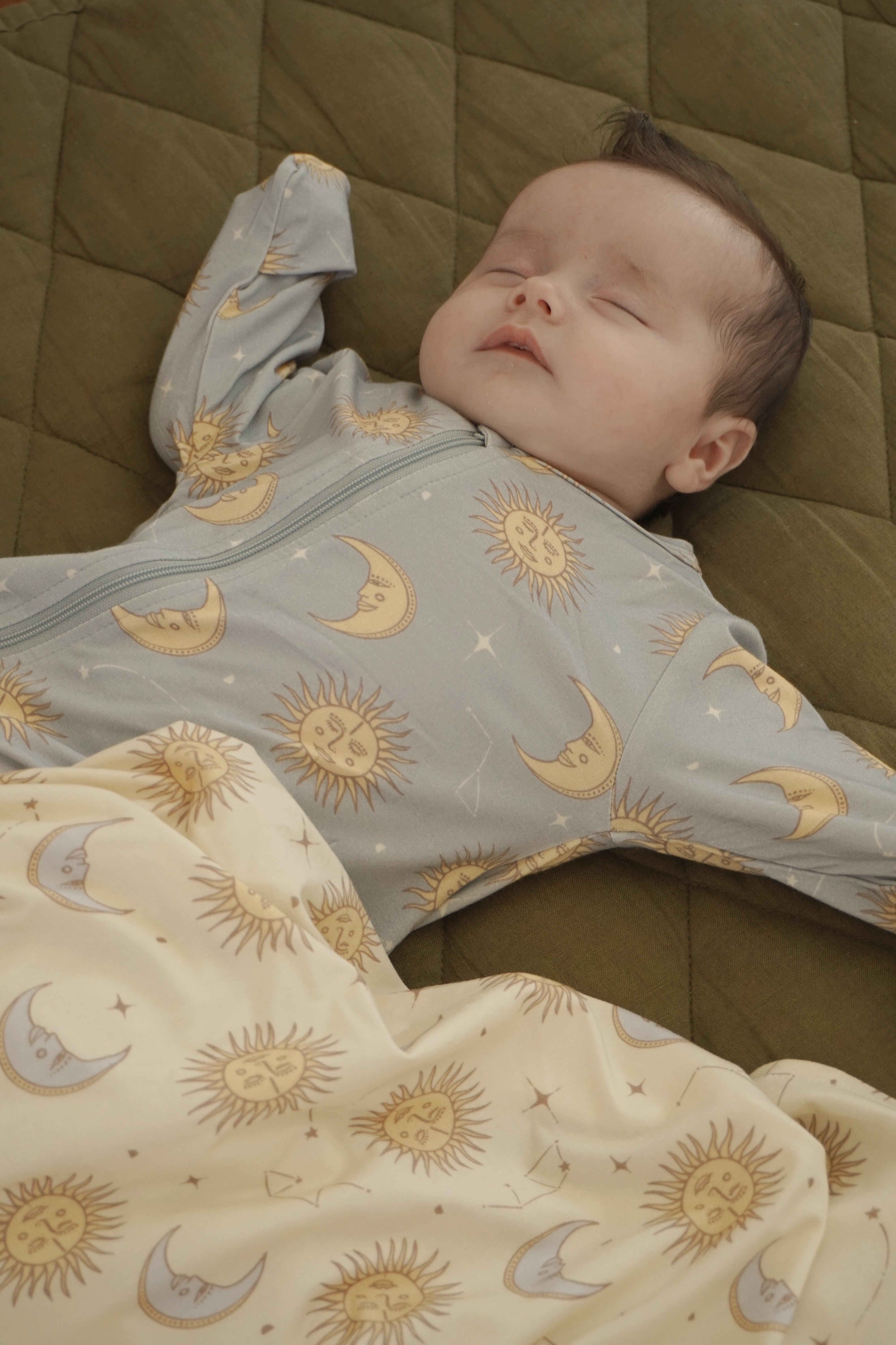 A sleeping baby in the Celestial Zip Jumpsuit Dusty Blue by BAM LOVES BOO lies on a green quilt. The baby, cozy in the bamboo organic cotton outfit, is partially covered with a cream-colored blanket featuring matching sun and moon designs.