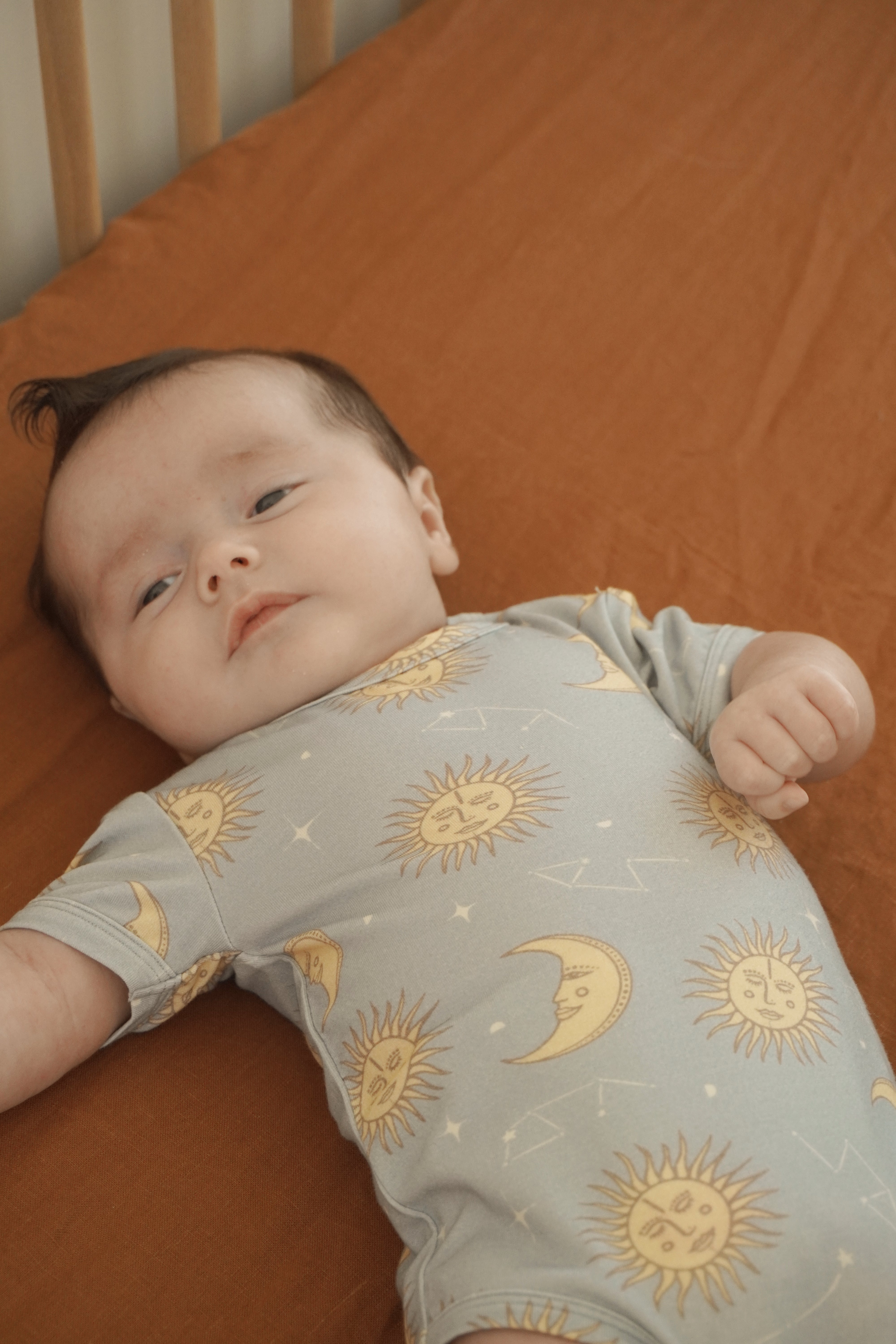 A baby lies on a brown bed, dressed in the BAM LOVES BOO Celestial Short Sleeved Onesie Dusty Blue. The onesie, made from bamboo organic cotton, features celestial print designs. The baby appears content and is gazing upward with one hand slightly curled.