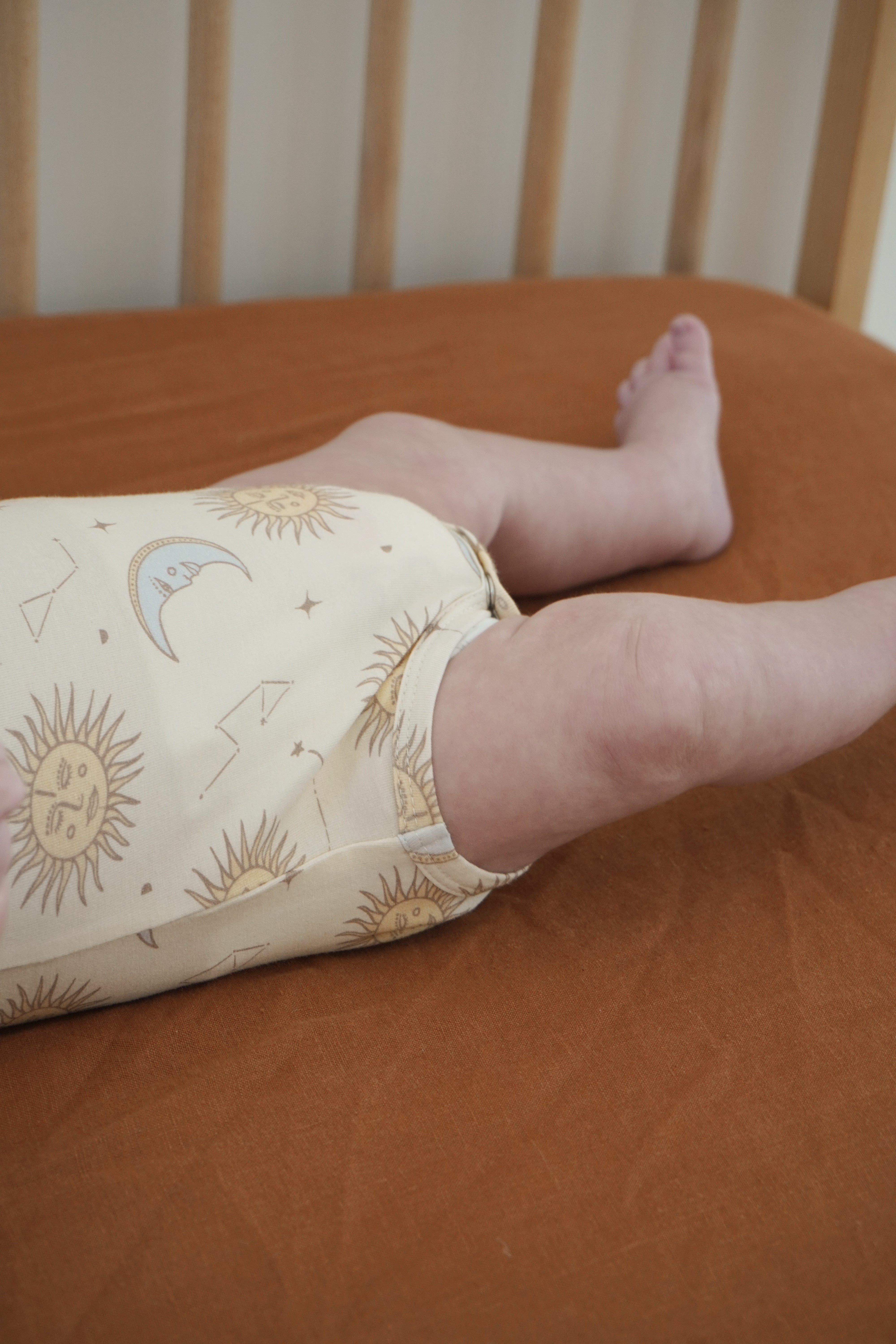 A baby lies on a brown sheet in a crib, dressed in a cream-colored Celestial Short Sleeved Onesie by BAM LOVES BOO, featuring an enchanting print of suns and moons. This outfit, made from bamboo organic cotton with eco-friendly dyes, showcases just its adorable legs.