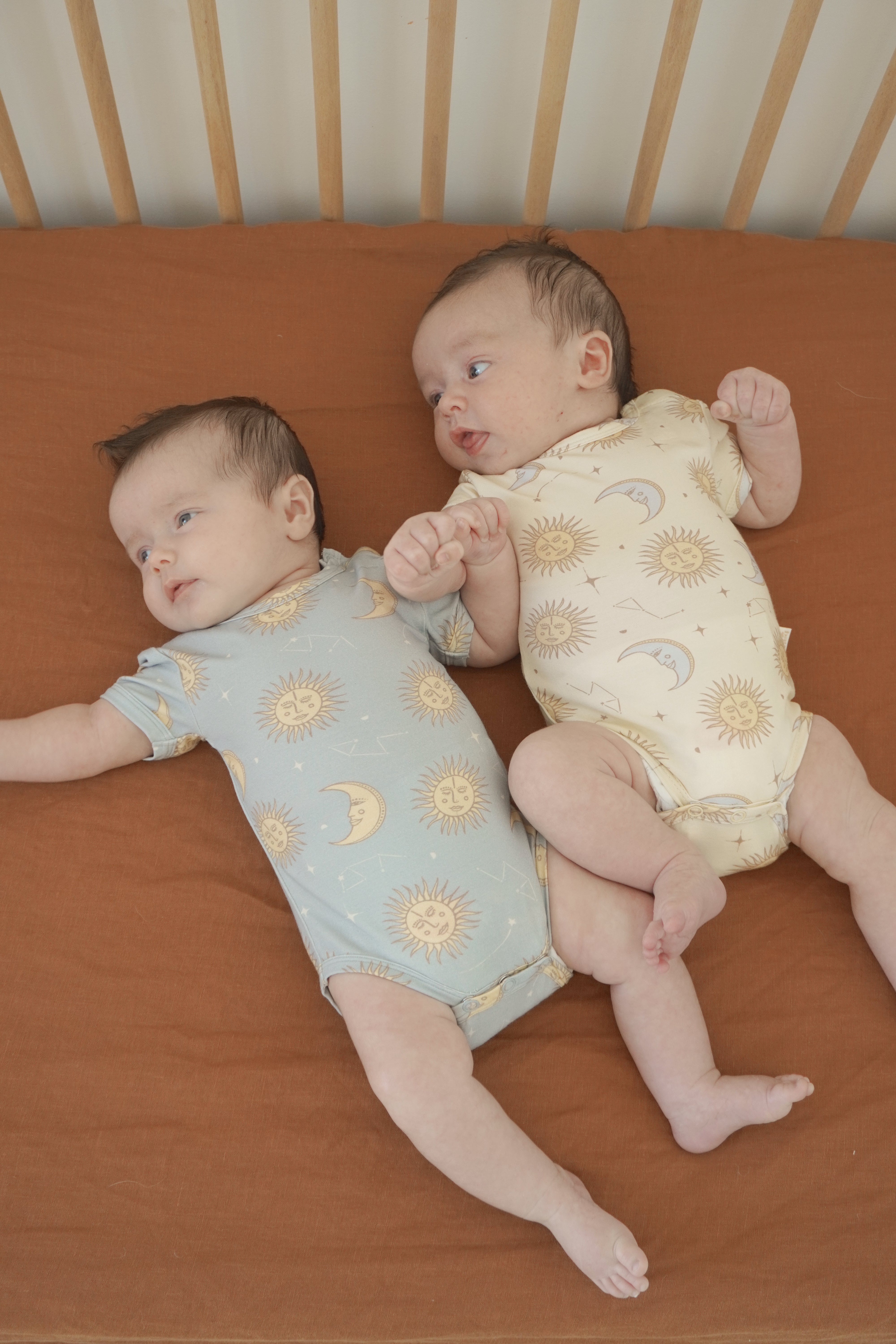 Two babies rest on a brown blanket in a crib, wearing matching BAM LOVES BOO onesies crafted from bamboo organic cotton and eco-friendly dyes. The celestial print showcases sun and moon patterns, with one in blue and the other in the Celestial Short Sleeved Onesie Cream. Their hands touch gently as they gaze in different directions.