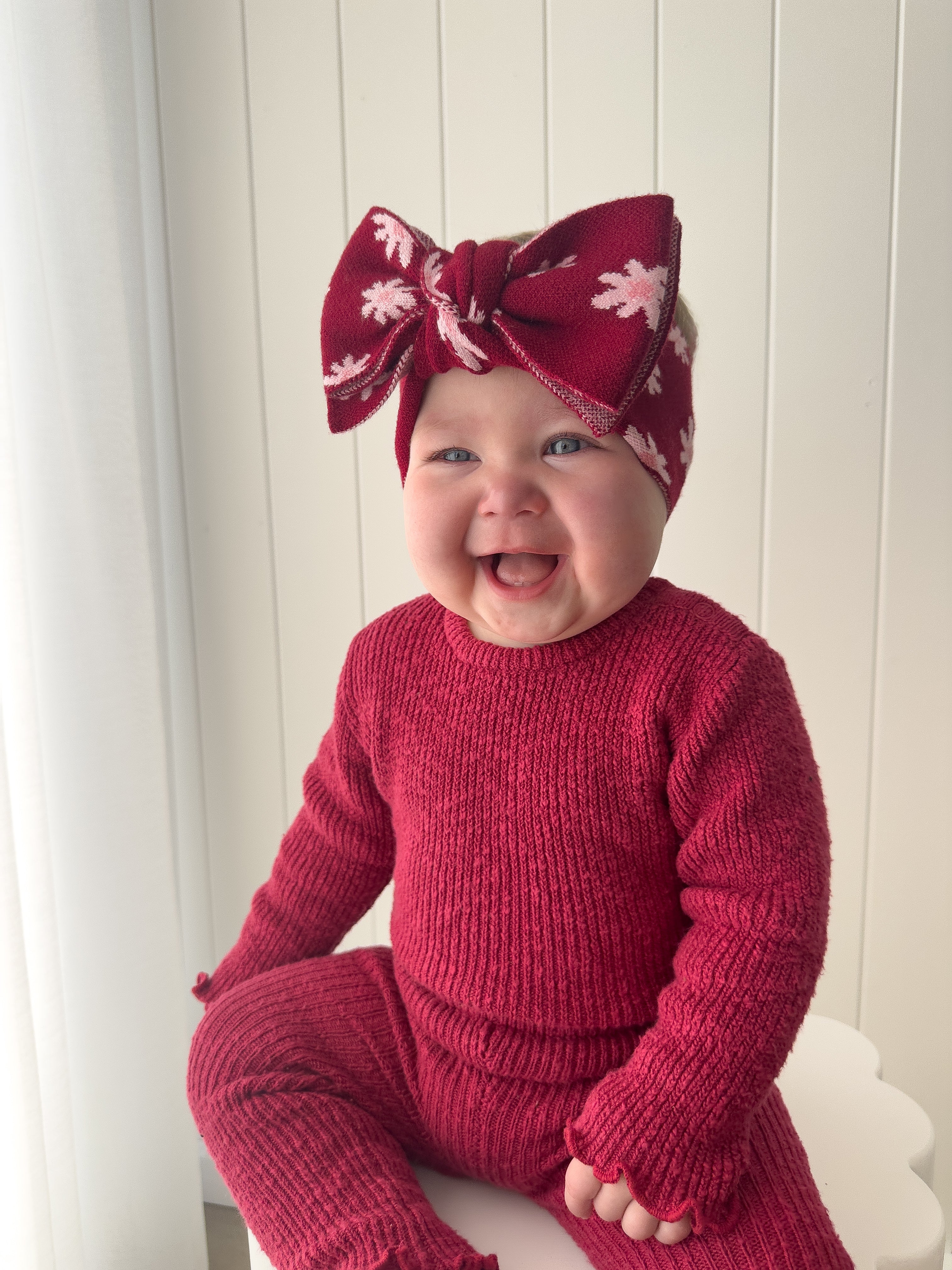 A smiling baby wears a ZIGGY LOU Ziggy Lou - Headband Lola (Plum), featuring a big bow, while sitting against a light paneled background.