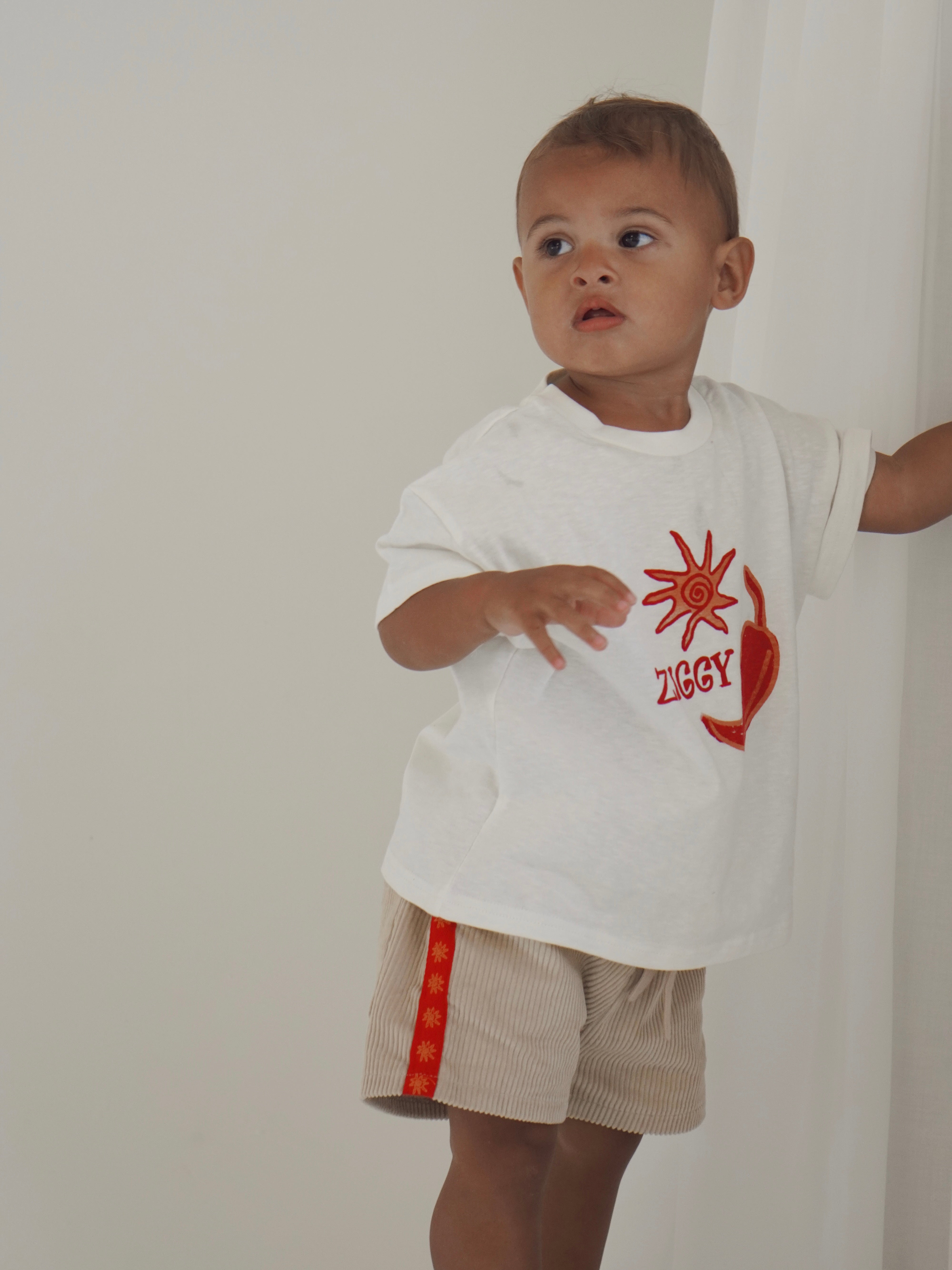 A toddler in a white t-shirt with a sun design and "Ziggy" stands by a curtain, showcasing Ziggy Lou's charm. They wear ZIGGY LOU's Cord Shorts Chilli—beige corduroy with bold red stripes—against a plain white wall.