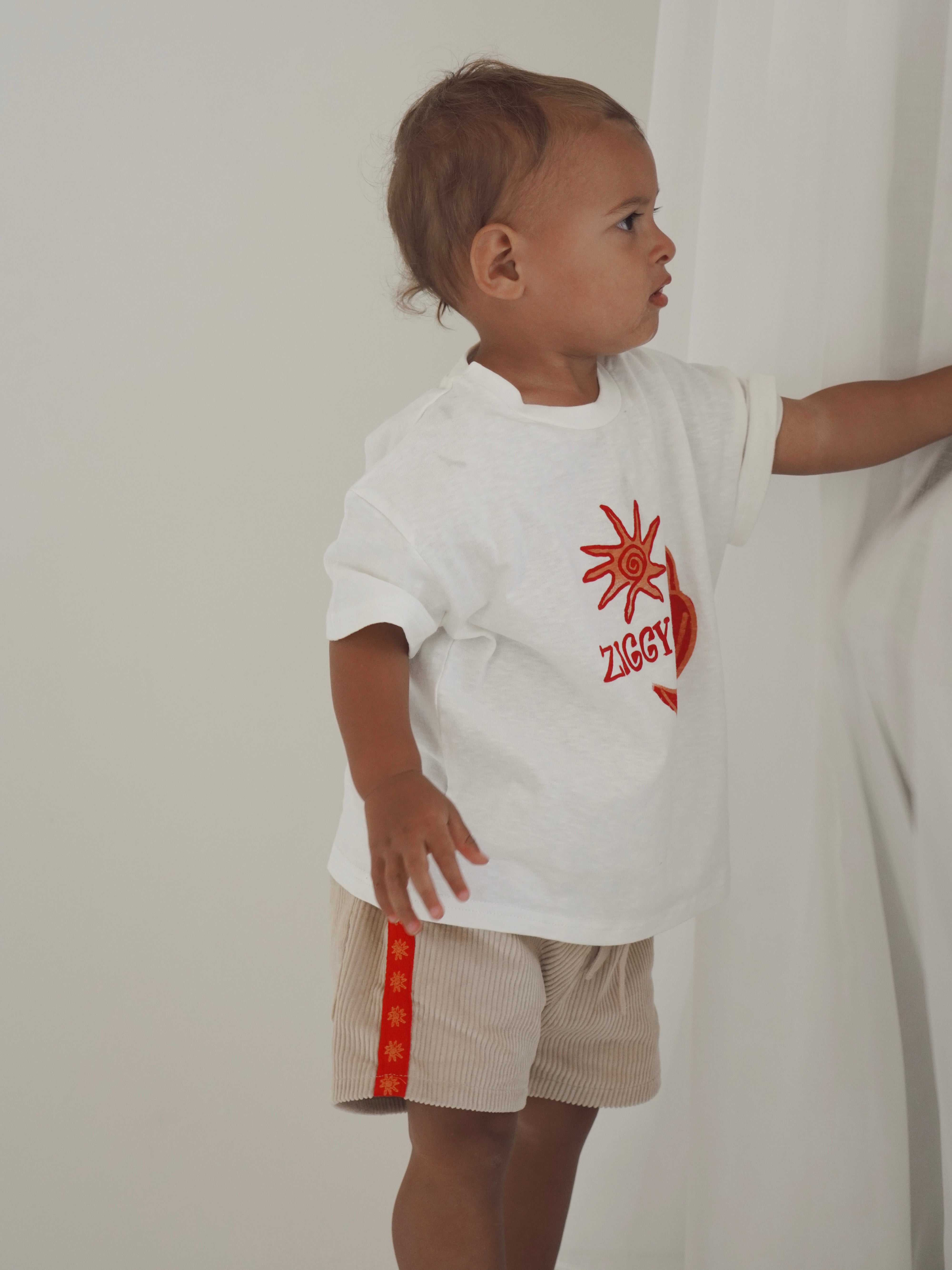 A toddler wearing a white T-shirt with a red sun graphic and the word "Ziggy" stands by a white curtain, gazing to the side. They sport ZIGGY LOU's Ziggy Lou ~ Cord Shorts Chilli, beige cotton corduroy shorts with a red side stripe, showcasing playful style.