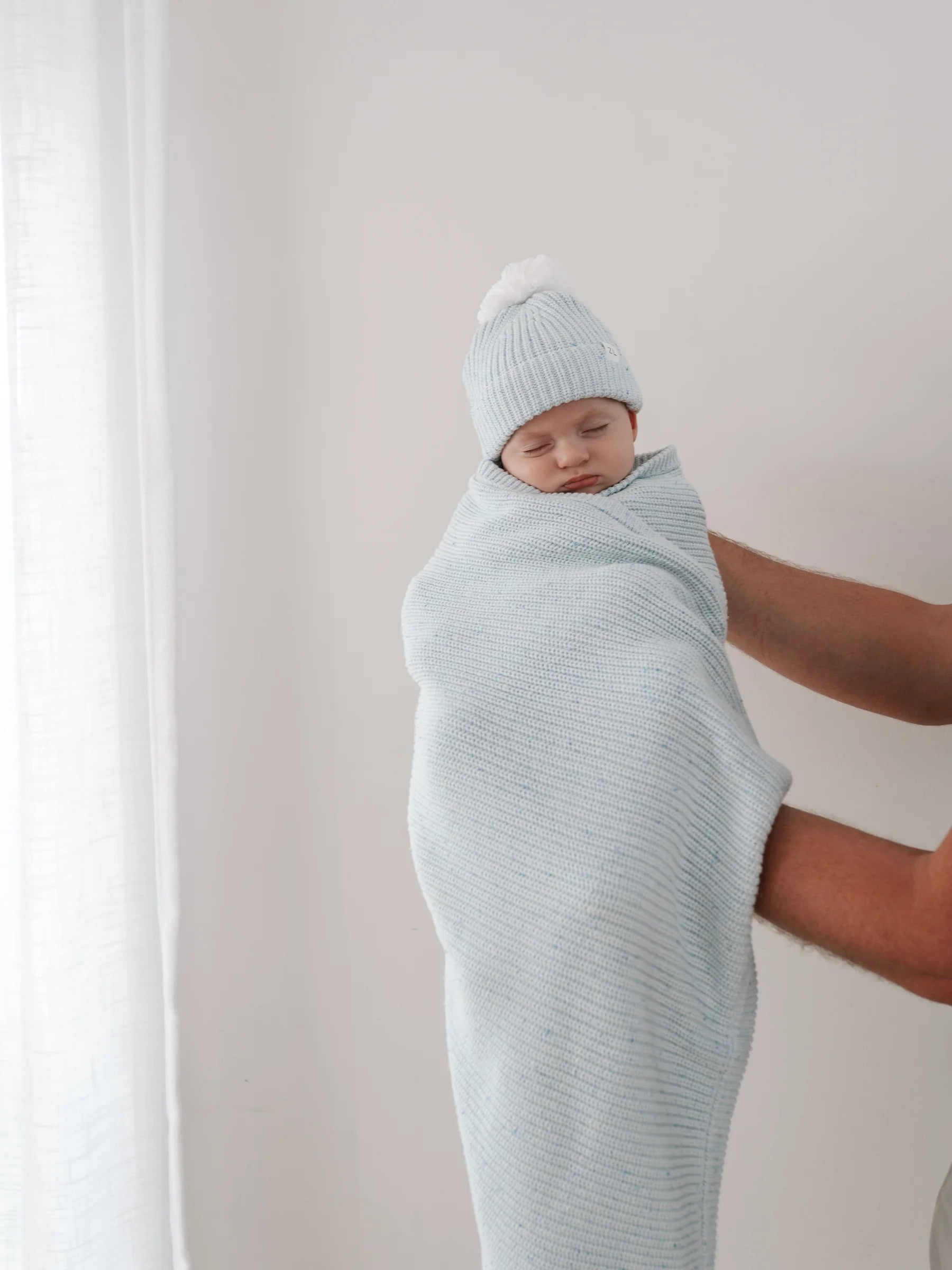 A baby sleeps wrapped in the ZIGGY LOU Ziggy Lou - Blanket Aero Fleck—crafted from 100% cotton in soothing Aero Fleck colour—with a matching hat, gently held in someone's arms against a white background.