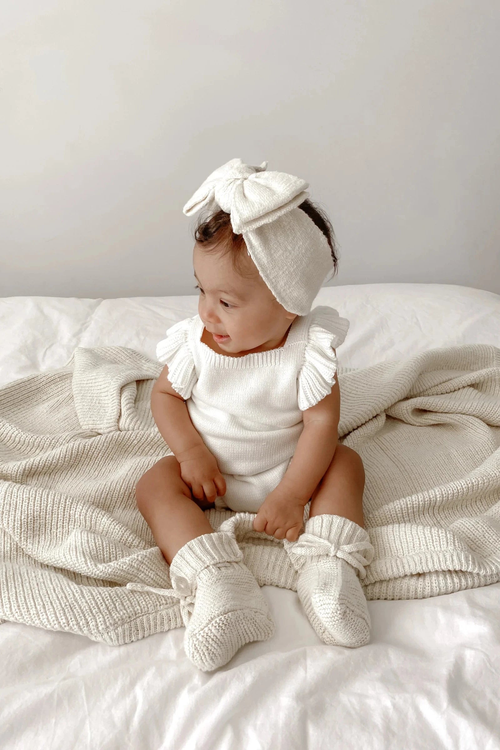 A baby is sitting on a bed covered with a knitted blanket. The baby is wearing a white outfit with ruffled sleeves, matching knit socks, and 100% textured cotton Booties Honey by ZIGGY LOU. A large white headband with a bow completes the look as the baby gazes to the side with a cheerful expression.