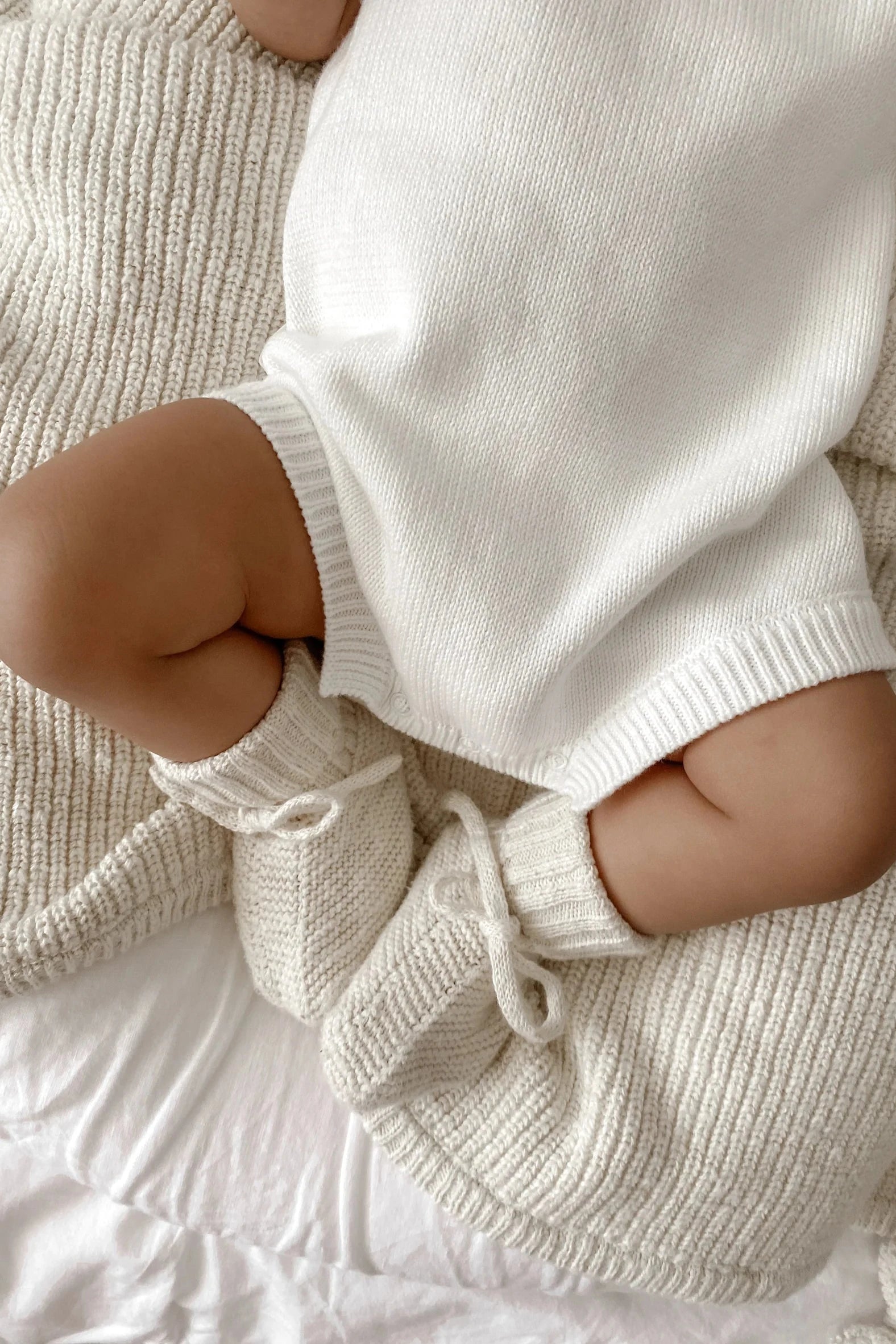 A baby wearing a white knitted outfit and ZIGGY LOU Booties Honey, made from 100% textured cotton, sits on a cozy knitted blanket. The soft textures in the image create a warm, comforting atmosphere. The baby's chubby legs and gentle attire highlight the serene and tender moment, making it feel like an heirloom piece.