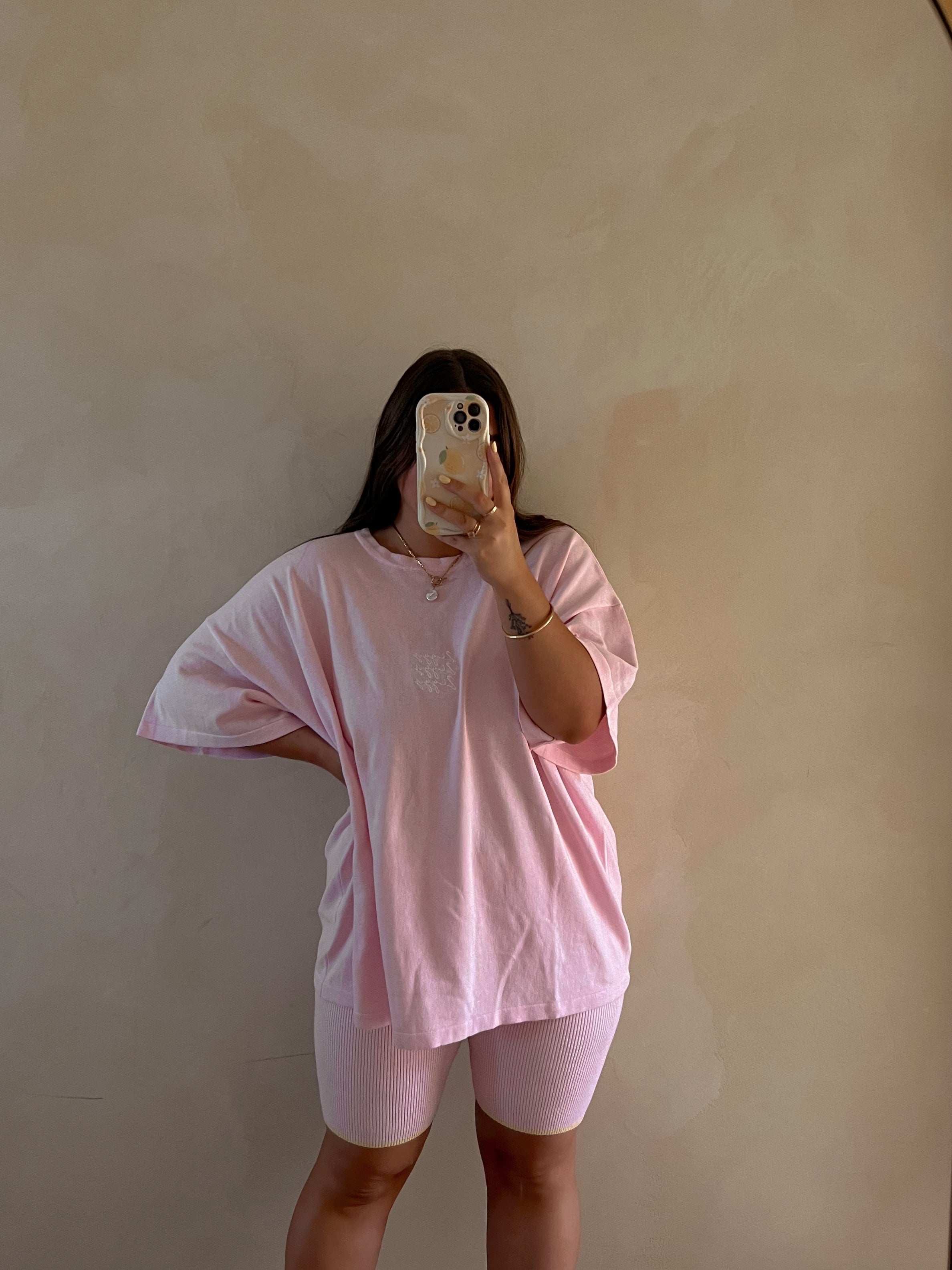 Wearing the Women's Signature Tee Freya by ZIGGY LOU in an oversized pink style, paired with ribbed shorts, a person takes a mirror selfie with a mobile phone against a light beige background, showing only their silhouette and outfit.