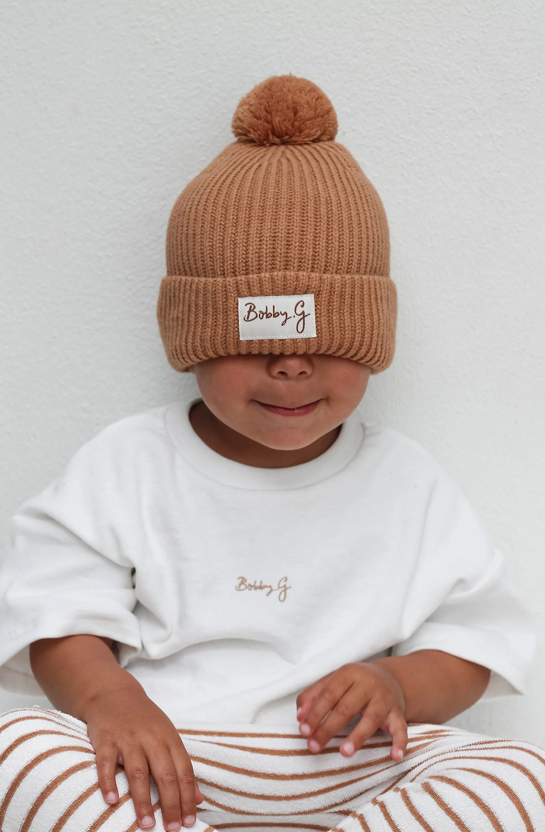 A young toddler wearing the Camden caramel striped pants with a white shirt and the pom pom beanie in rusty. The beanie is pulled over their eyes.