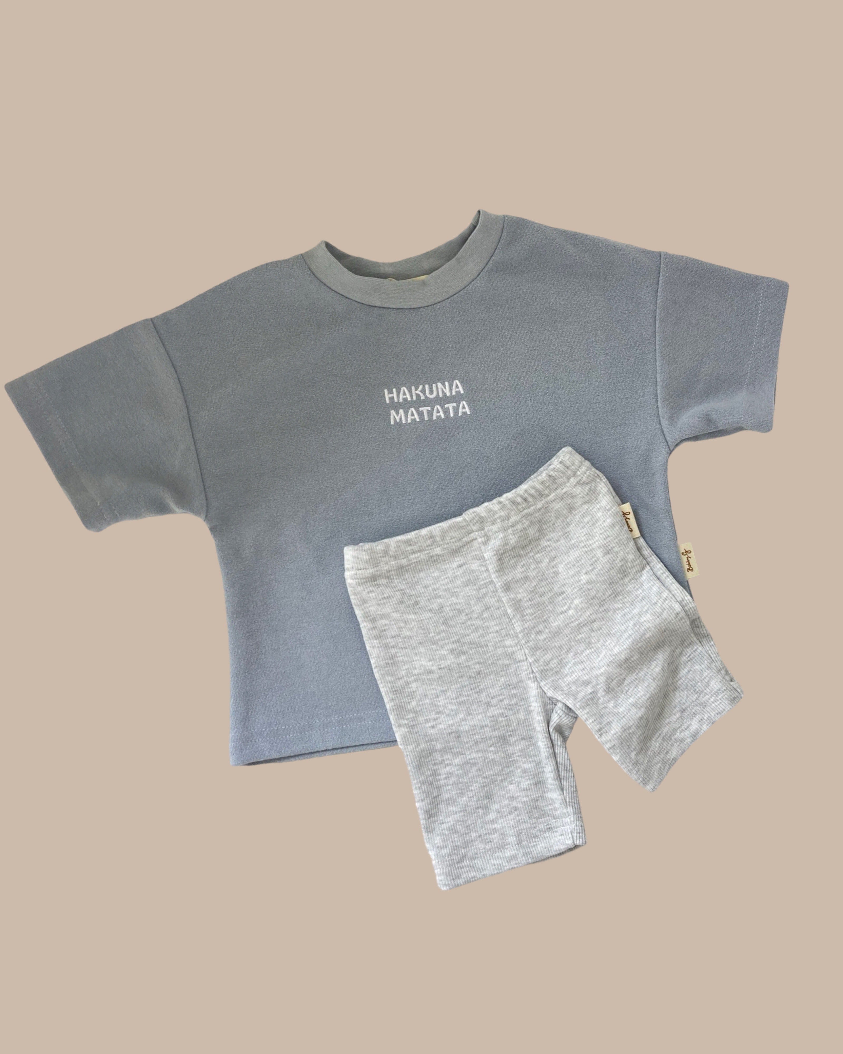 A child's outfit is presented against a beige background, ideal for summer essentials. The set features a light blue short-sleeved top with "Hakuna Matata" on the front, matched with BOBBY G BABY WEAR's Bike Shorts Smoke in light gray – a delightful addition to any kids' wardrobe.