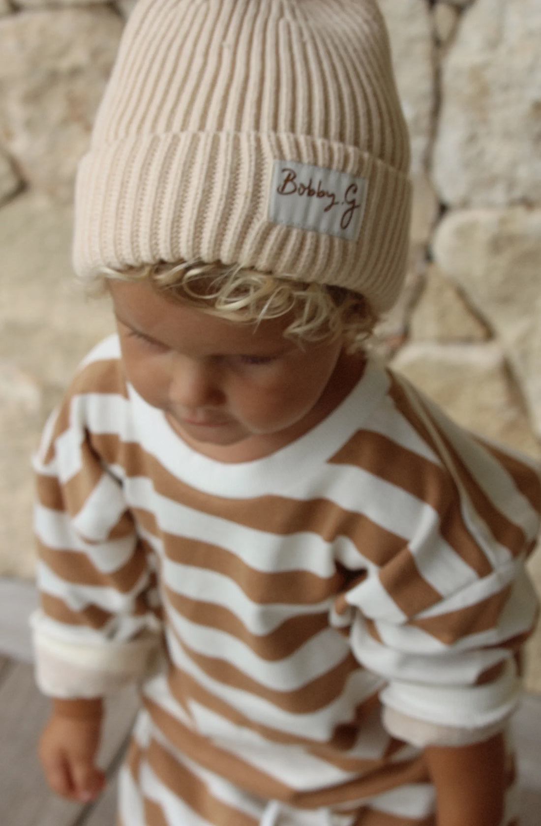 A young toddler wearing the Camden caramel striped pullover and matching pants and the pom pom beanie in cream.