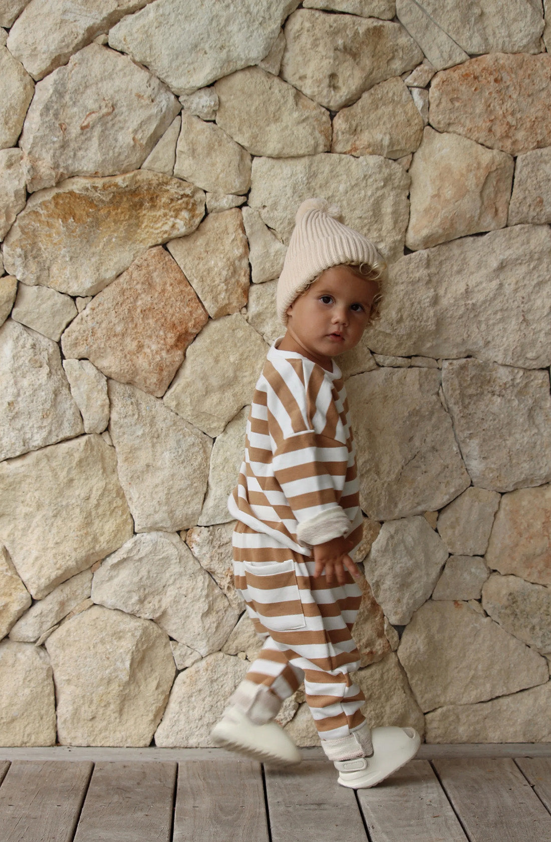 A young toddler wearing the Camden caramel striped pullover and matching pants with a puffer vest over the top. He is standing in front of a stone pebble wall. 